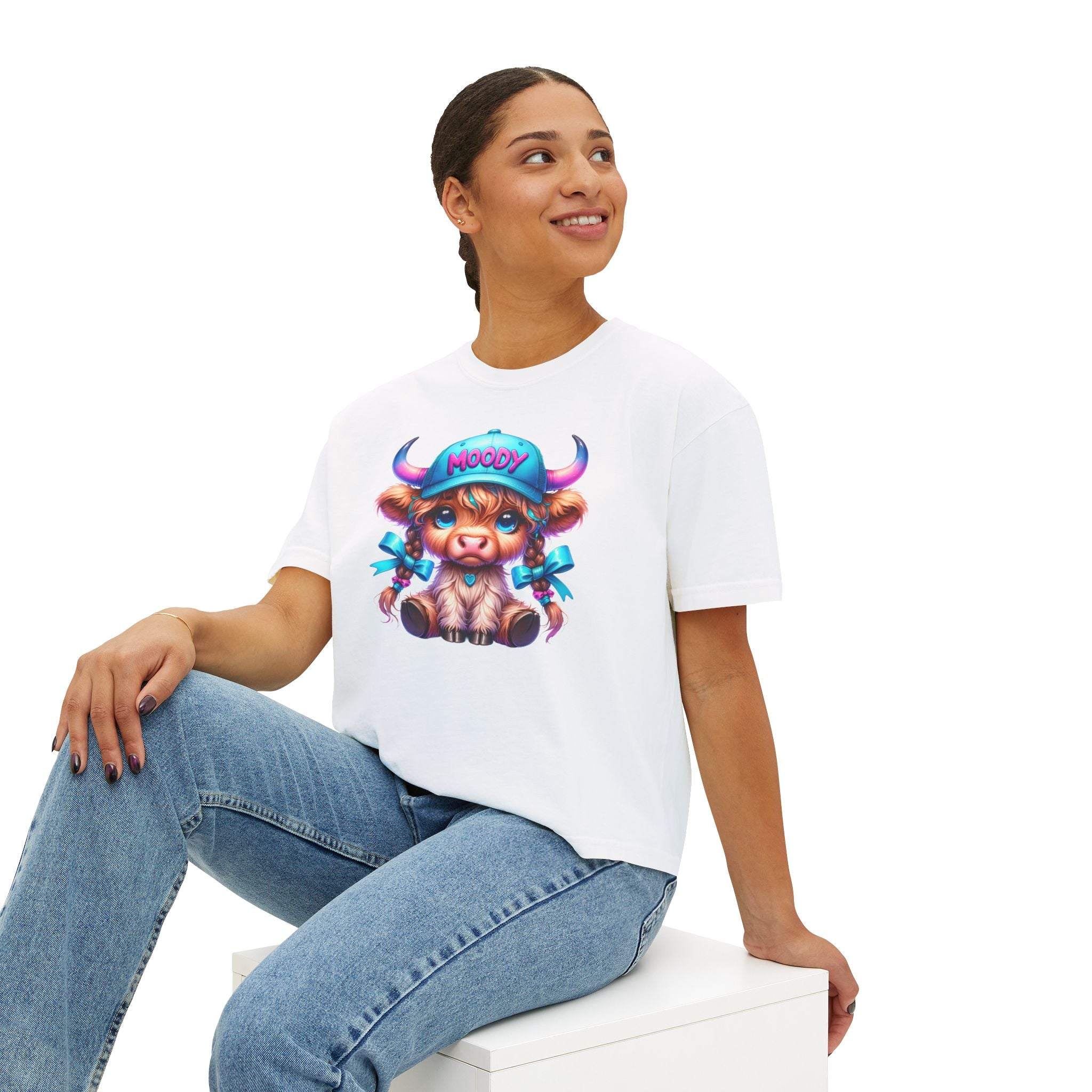 Woman wearing white boxy tee with moody girl highland cow graphic sitting on white block