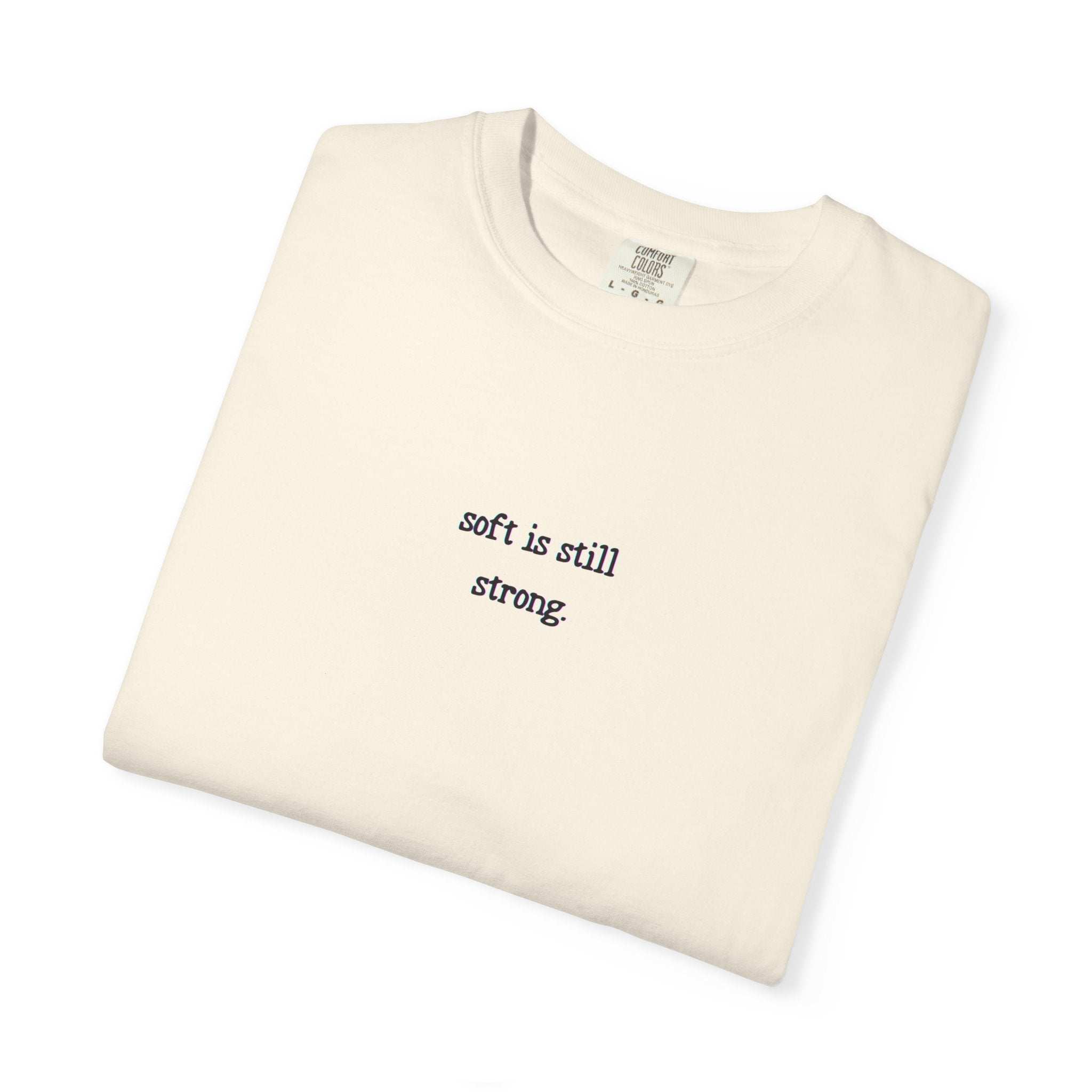 Folded cream-colored t-shirt with 'soft is still strong' text on a white background