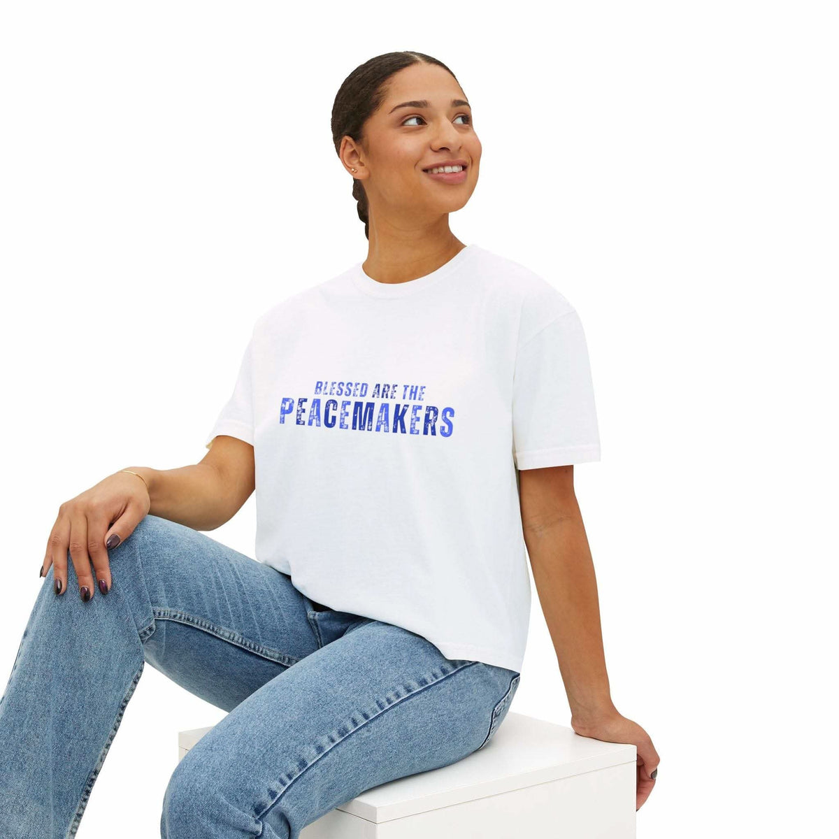 Woman wearing white Tee Blessed be the Peacemakers boxy shirt with blue text and jeans sitting on a white cube
