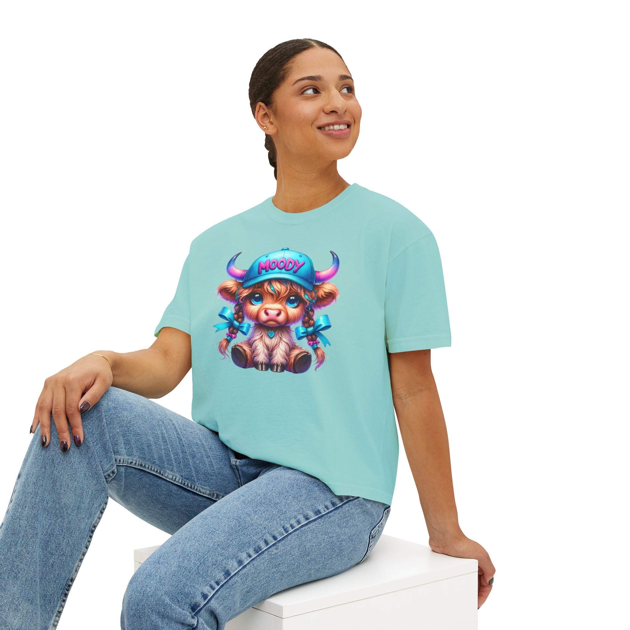 Woman wearing blue boxy tee with moody girl highland cow graphic sitting cross-legged