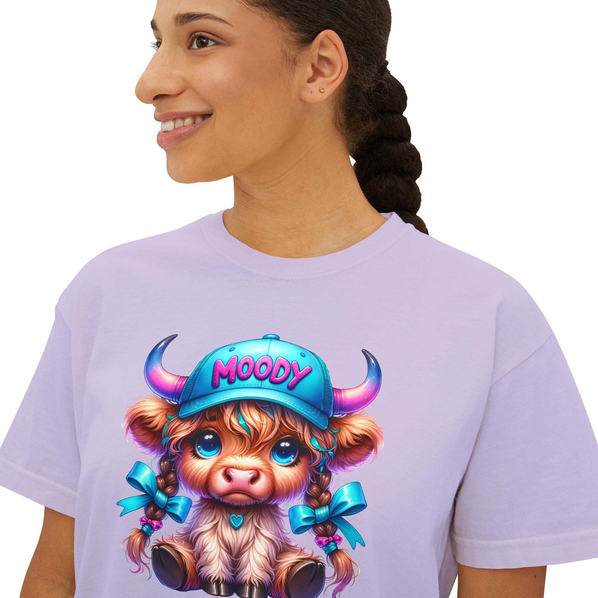 Woman wearing lavender boxy tee with moody girl highland cow graphic and blue cap