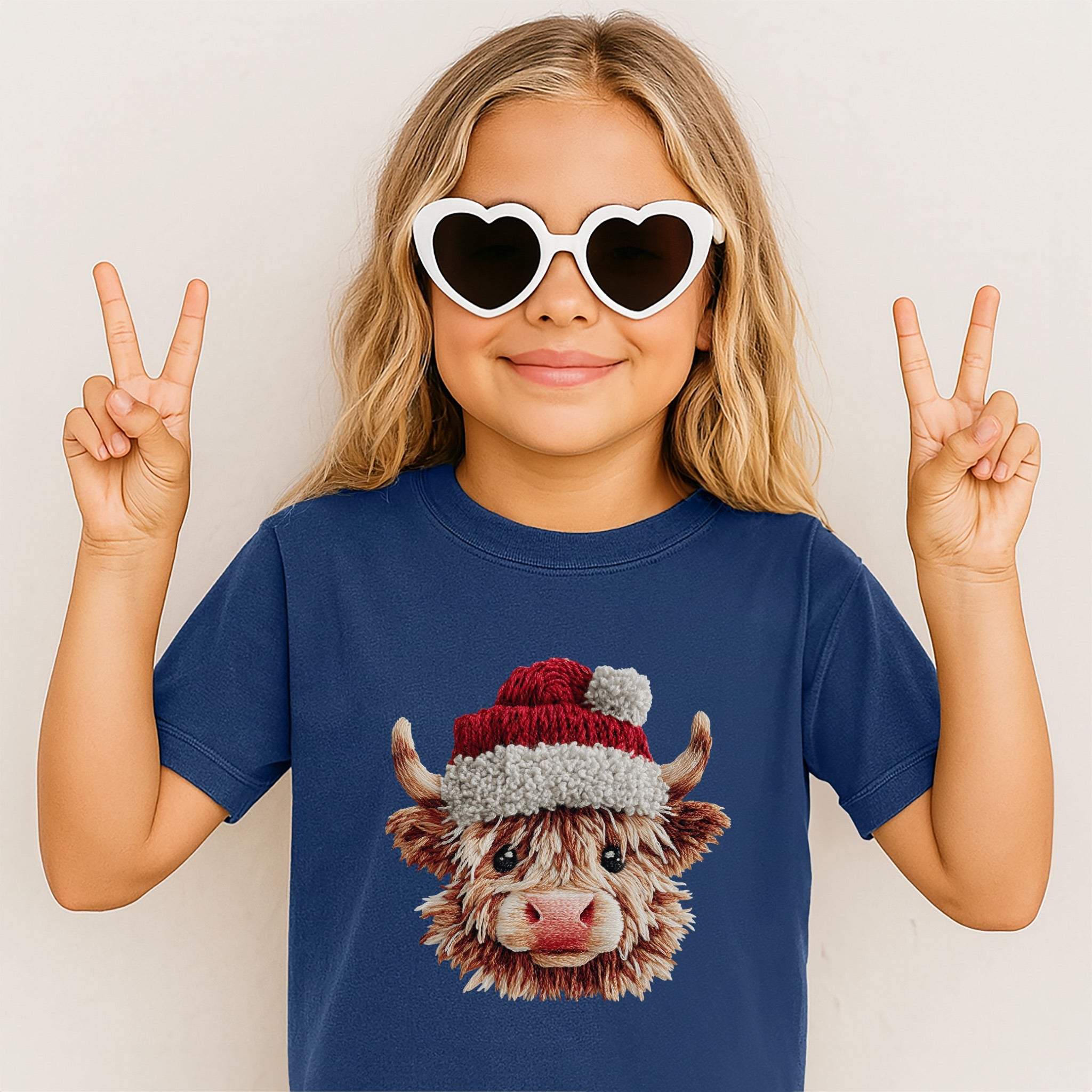 Child wearing navy Santa Highland Cow Youth Tee with faux crochet Highland cow in Santa hat design and heart-shaped sunglasses