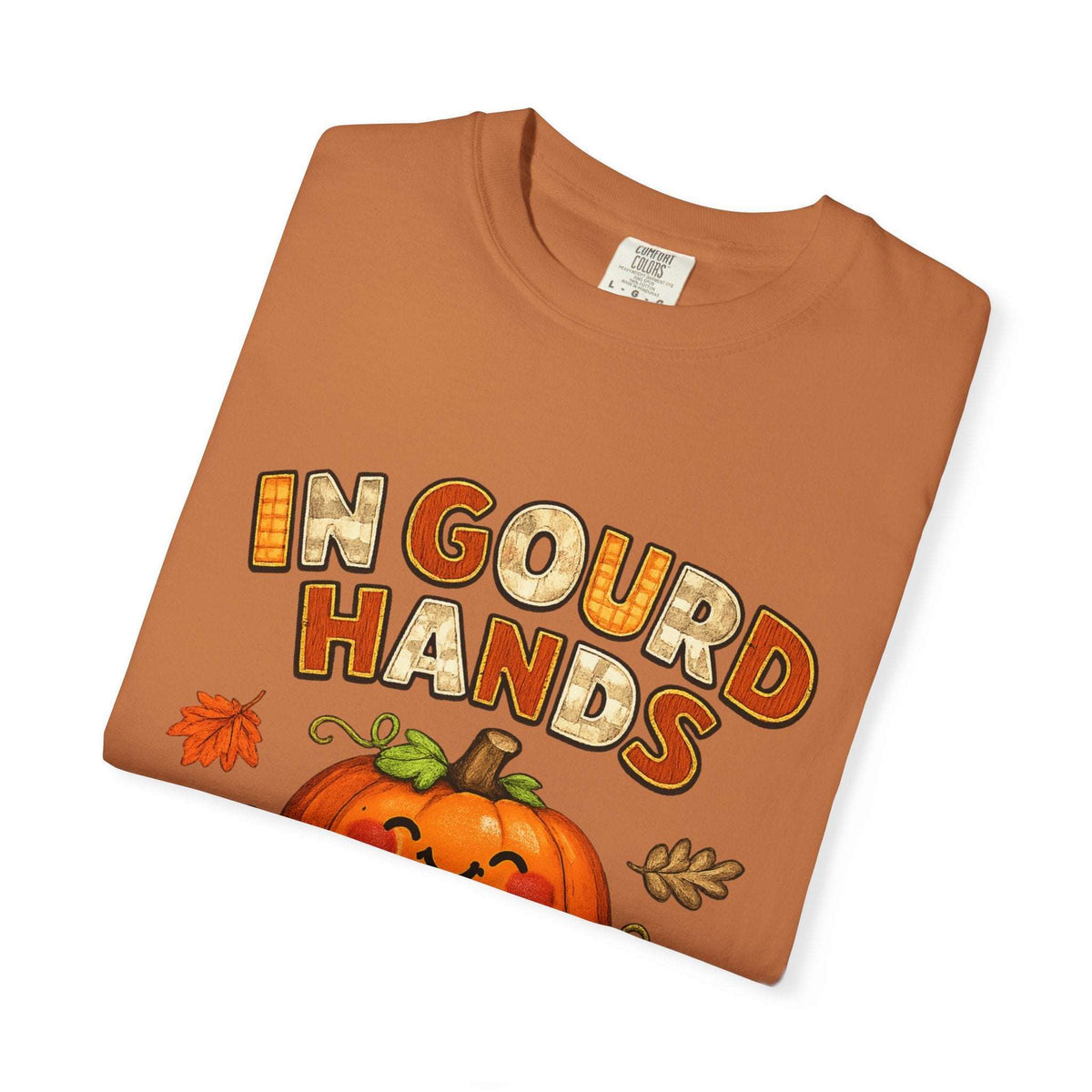 A folded orange crewneck t-shirt featuring a festive autumn graphic with the text 'IN GOURD HANDS' in a rustic orange and cream font, above a smiling jack-o'-lantern pumpkin and small fall leaves, shown on a plain white background.