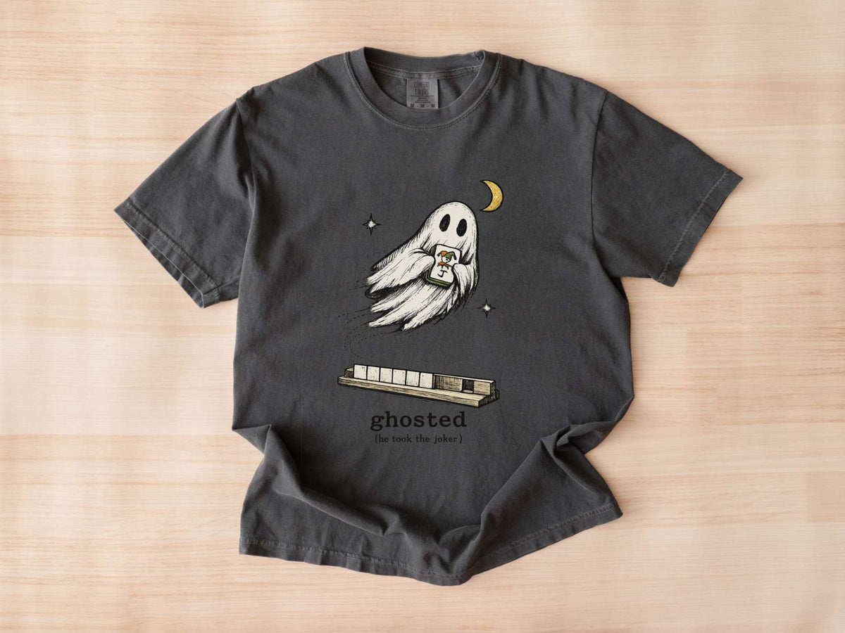 Flat lay of a pepper mahjong T-shirt with a ghost holding a joker tile above a tile rack and “ghosted (he took the joker)” lettering.