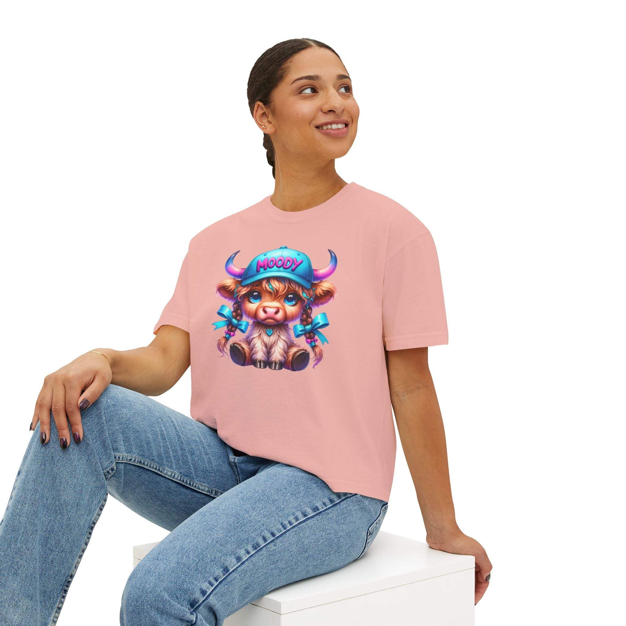 Woman wearing pink boxy tee with moody girl highland cow graphic sitting on white block