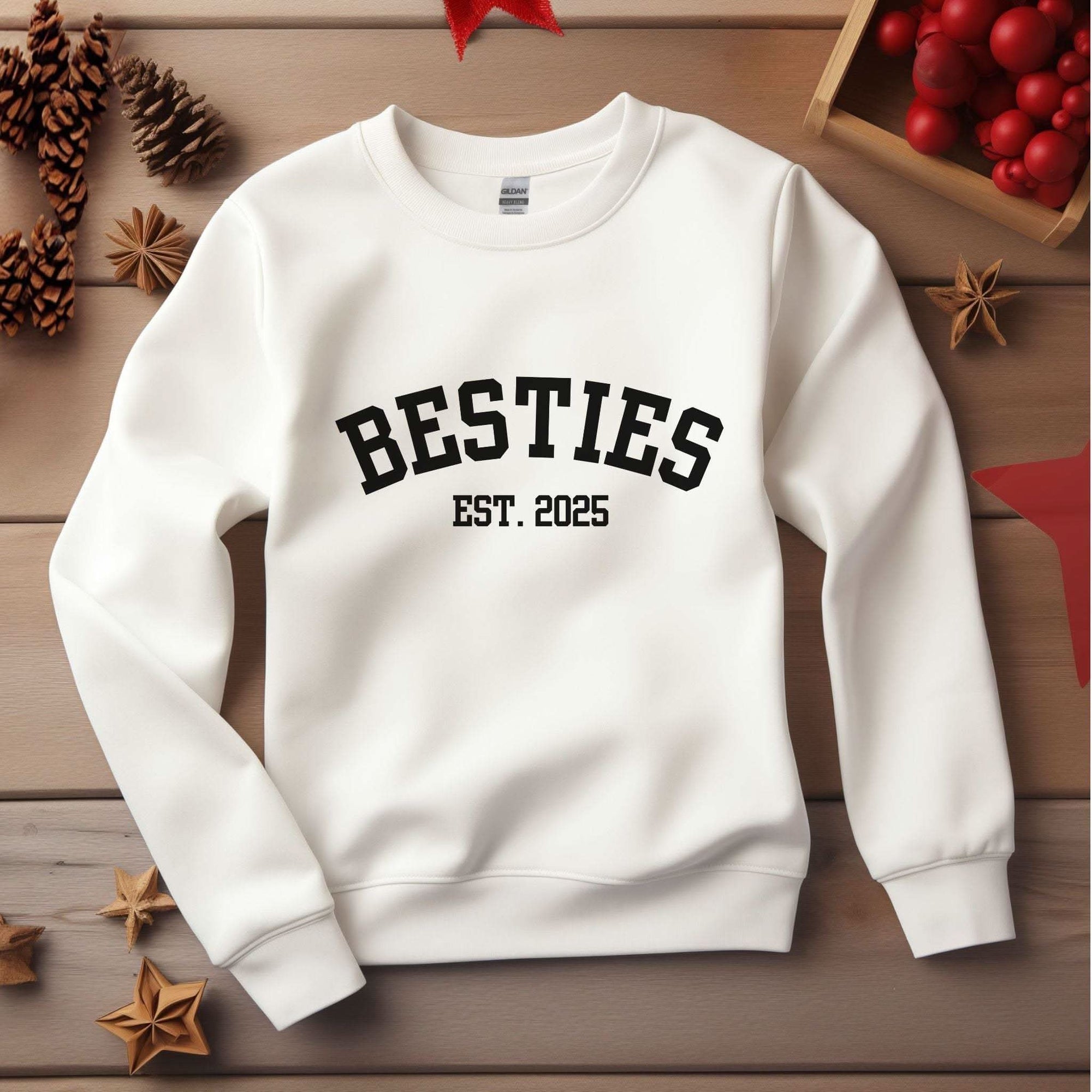 White Varsity Besties Crewneck Sweatshirt with black "BESTIES EST. 2025" lettering on a wooden background