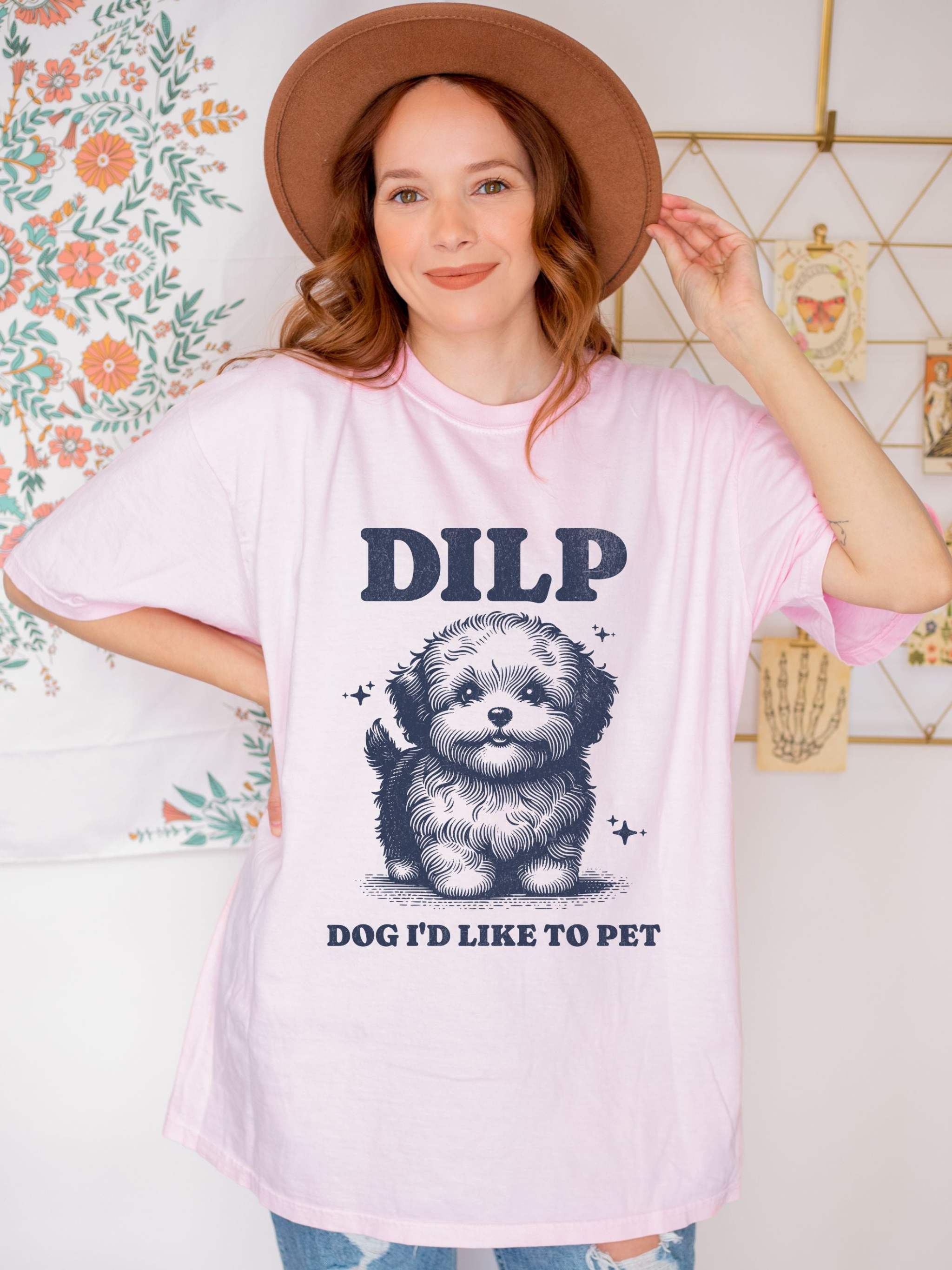 Garment-dyed pink T-shirt with cute dog graphic and "DILP Dog I'd Like To Pet" text worn by woman with brown hat