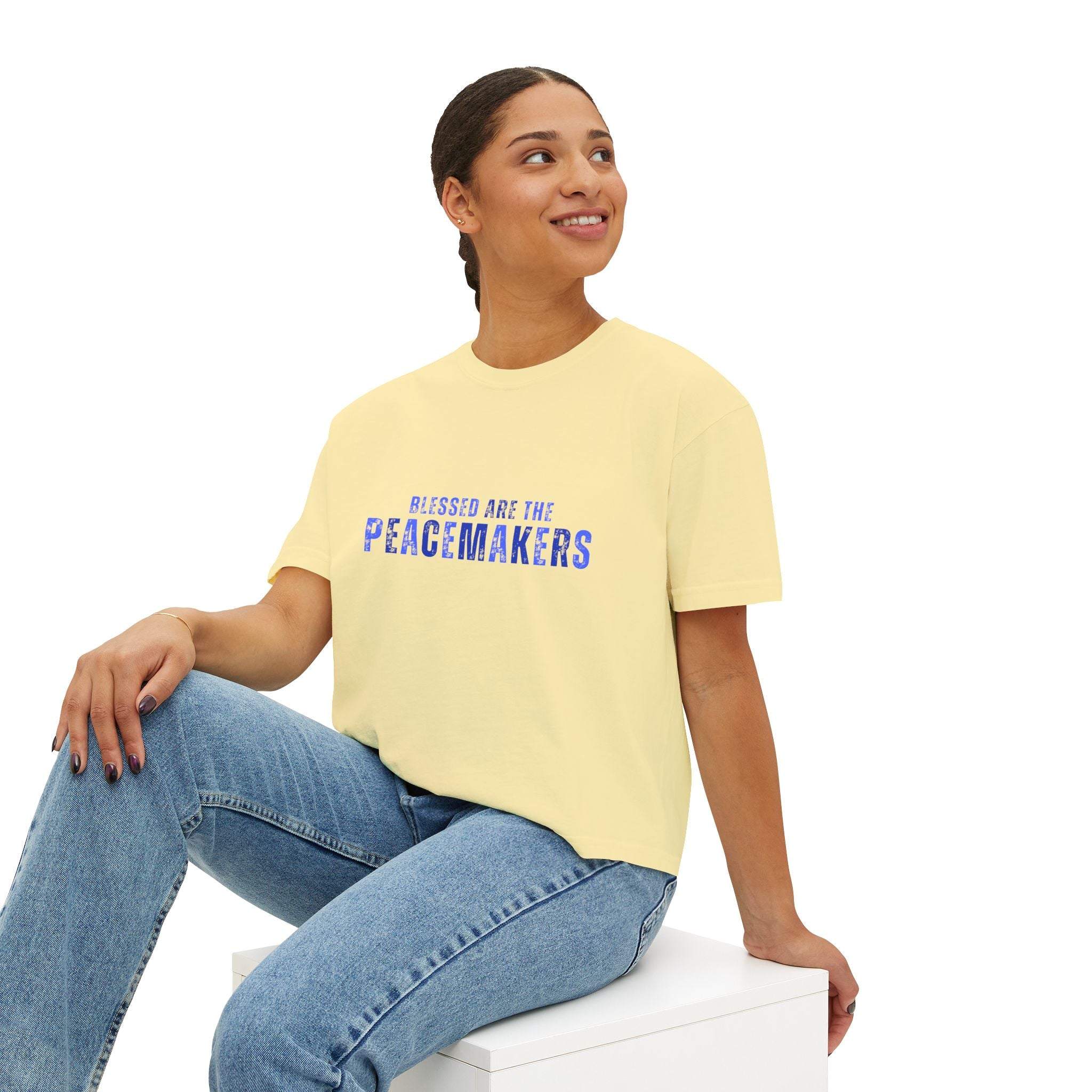 Woman wearing Tee Blessed be the Peacemakers boxy yellow shirt with blue text and jeans sitting on white block