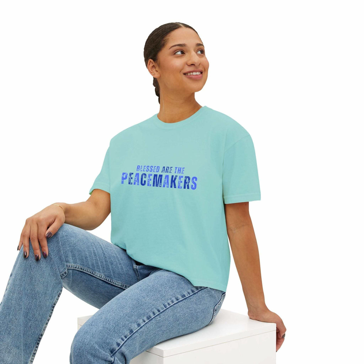 Woman wearing Tee Blessed be the Peacemakers boxy shirt in light blue with printed text and jeans sitting on white block