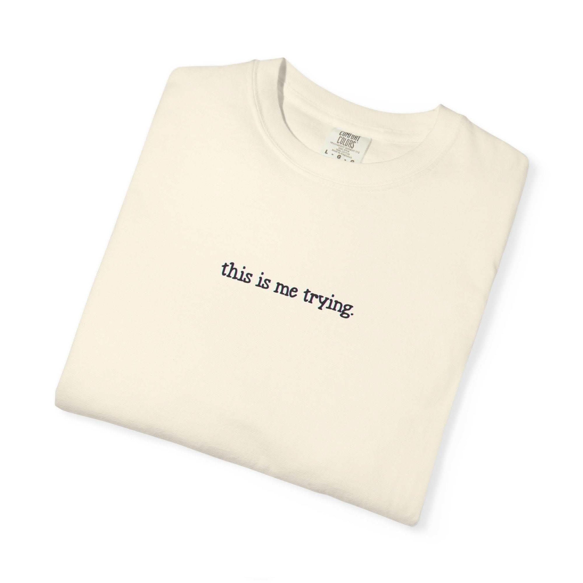 Folded beige t-shirt with 'this is me trying' text on a white background