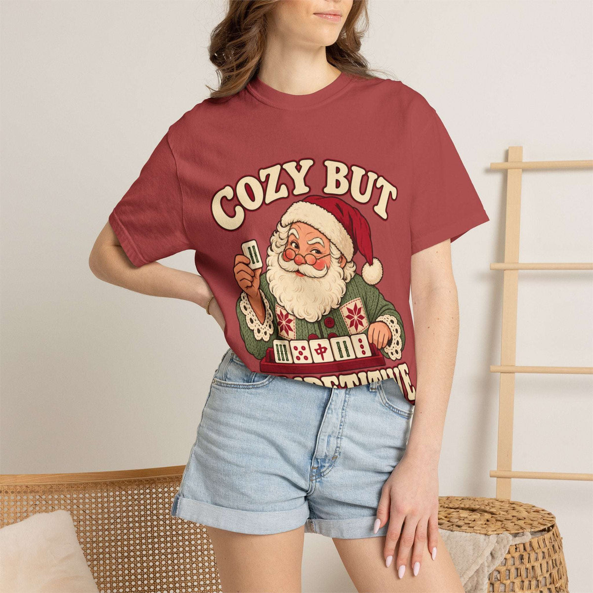 Model wearing a Crimson red Comfort Colors T-shirt with Santa holding a mahjong tile above a tile rack and cream “Cozy But Competitive” lettering.