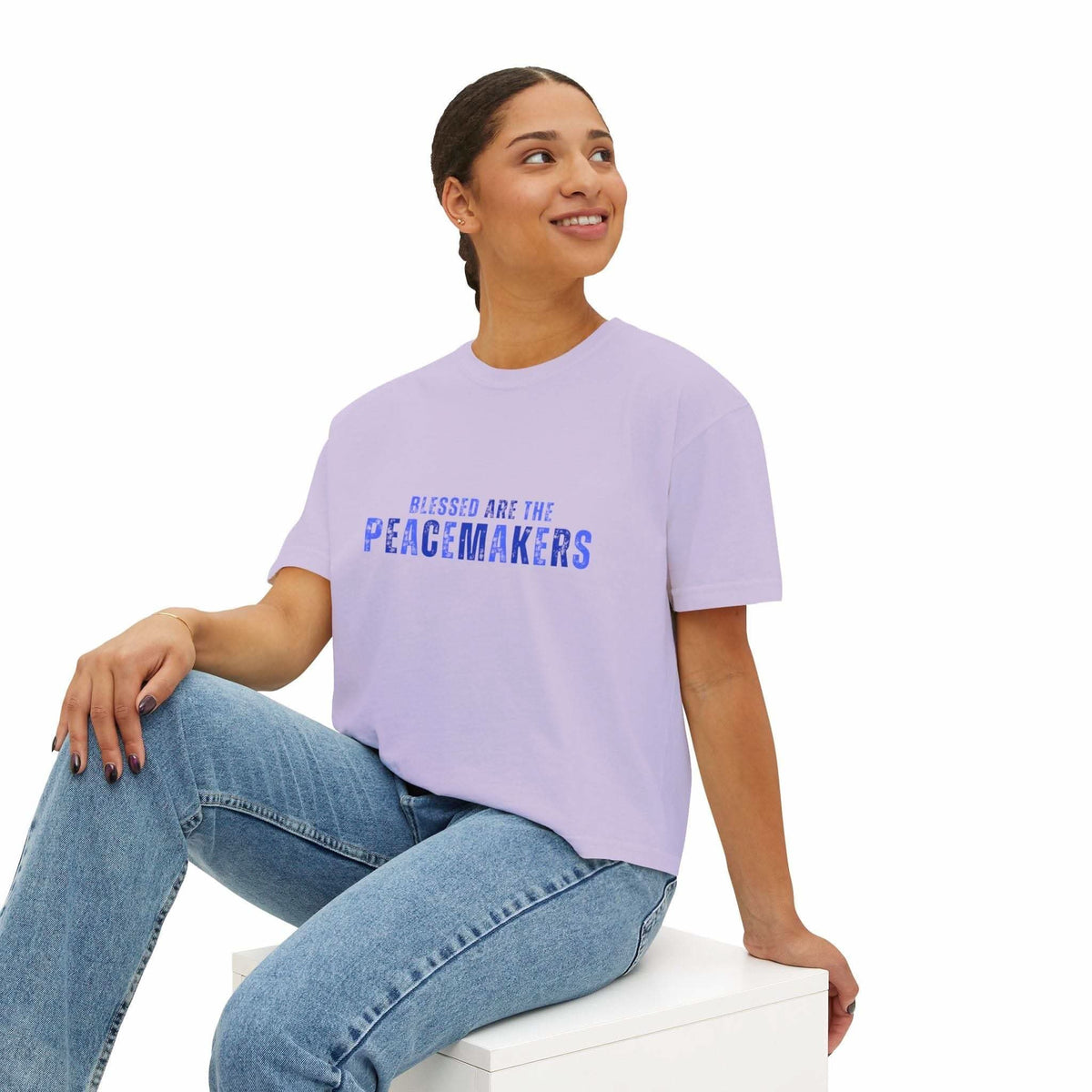 Woman wearing lavender Tee Blessed be the Peacemakers boxy shirt with blue text and jeans sitting on white block
