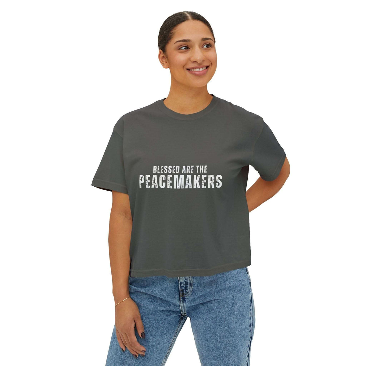 Woman wearing a dark gray Tee Blessed be the Peacemakers boxy shirt with white text and blue jeans