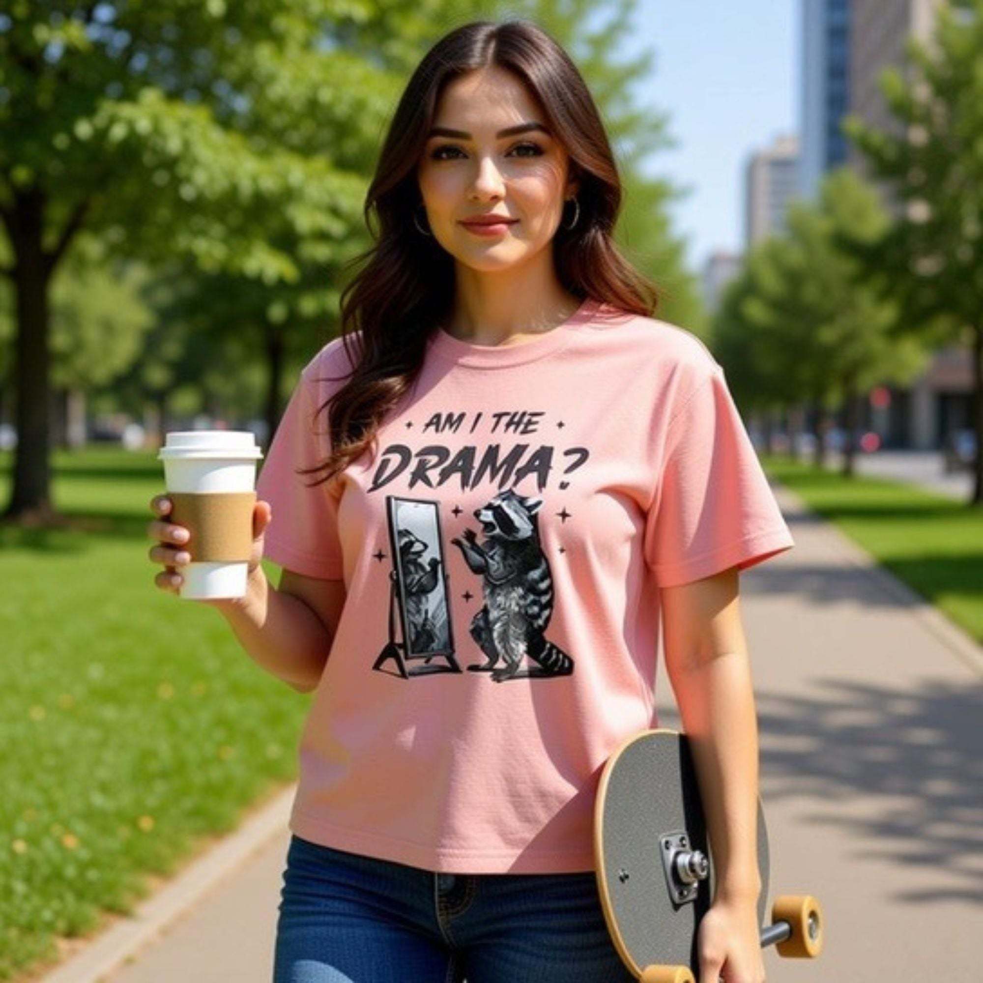 Woman wearing pink boxy tee with "Am I The Drama?" text and raccoon graphic, holding coffee and skateboard outdoors