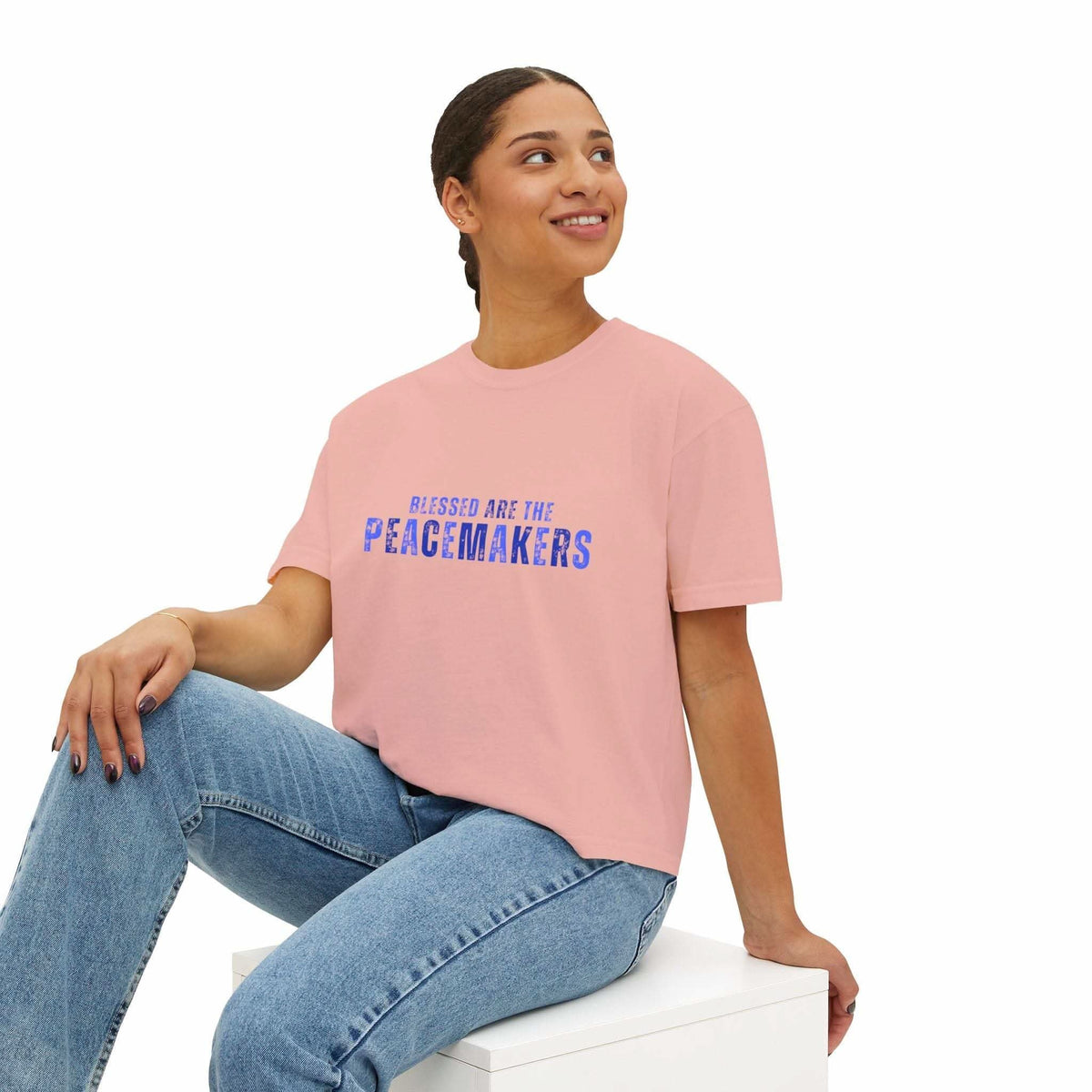 Woman wearing Tee Blessed be the Peacemakers pink boxy shirt with blue text and jeans sitting on white block