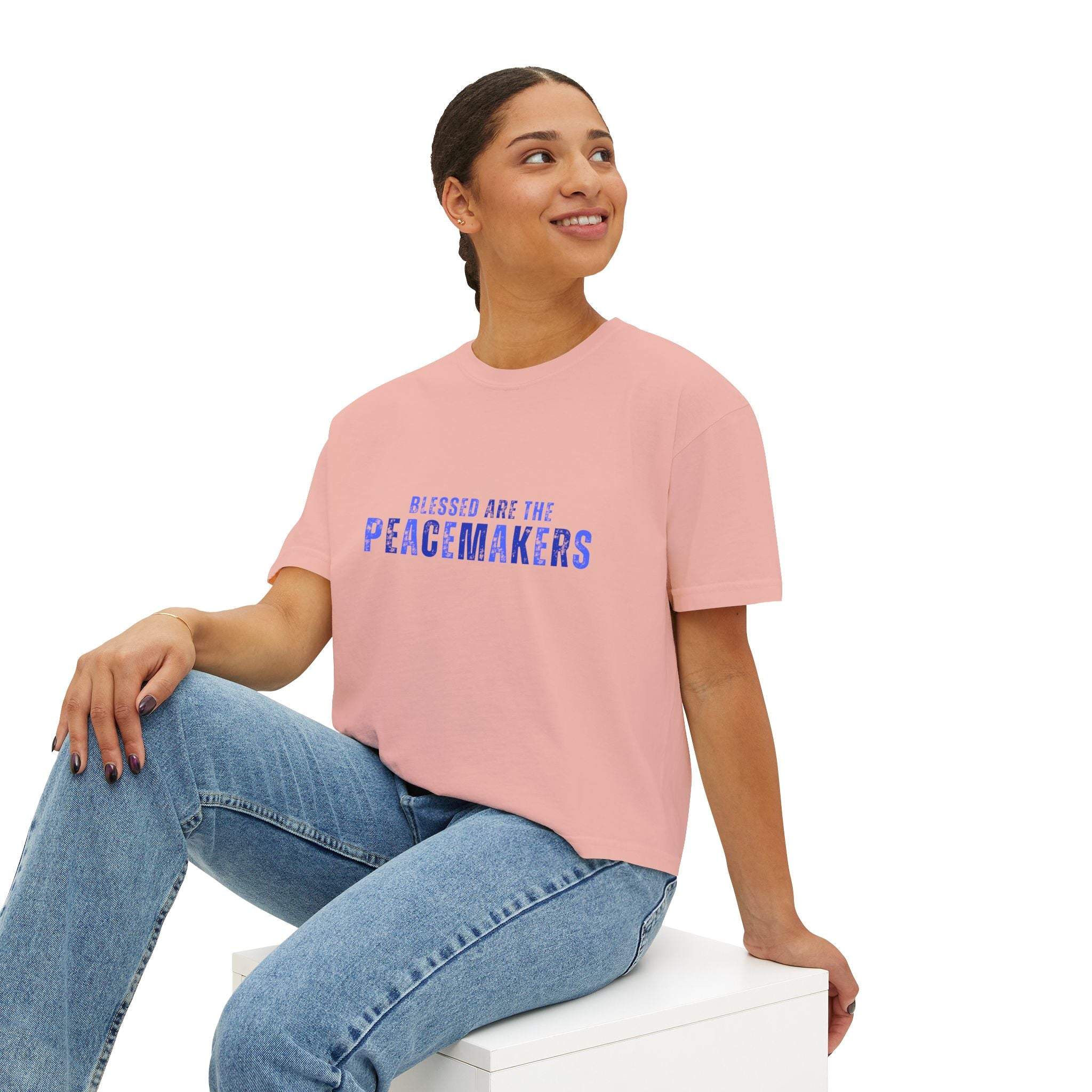 Woman wearing Tee Blessed be the Peacemakers pink boxy shirt with blue text and jeans sitting on white block
