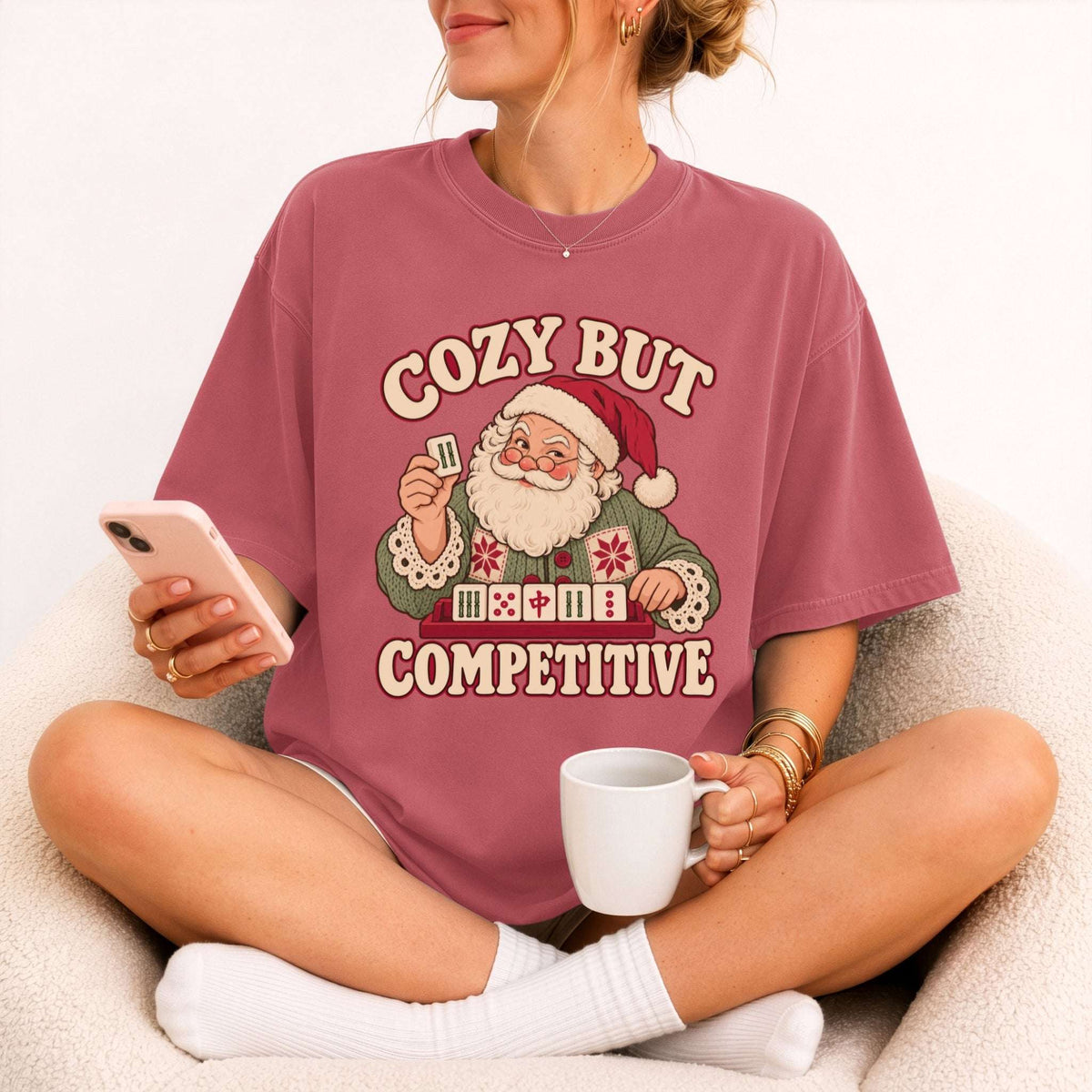 Model wearing a Crimson red Comfort Colors T-shirt with Santa holding a mahjong tile above a tile rack and cream “Cozy But Competitive” lettering.