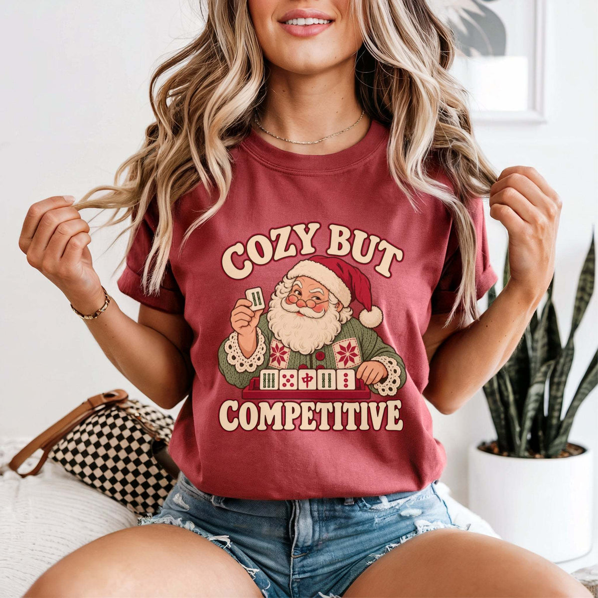 Model wearing a Crimson red Comfort Colors T-shirt with Santa holding a mahjong tile above a tile rack and cream “Cozy But Competitive” lettering.
