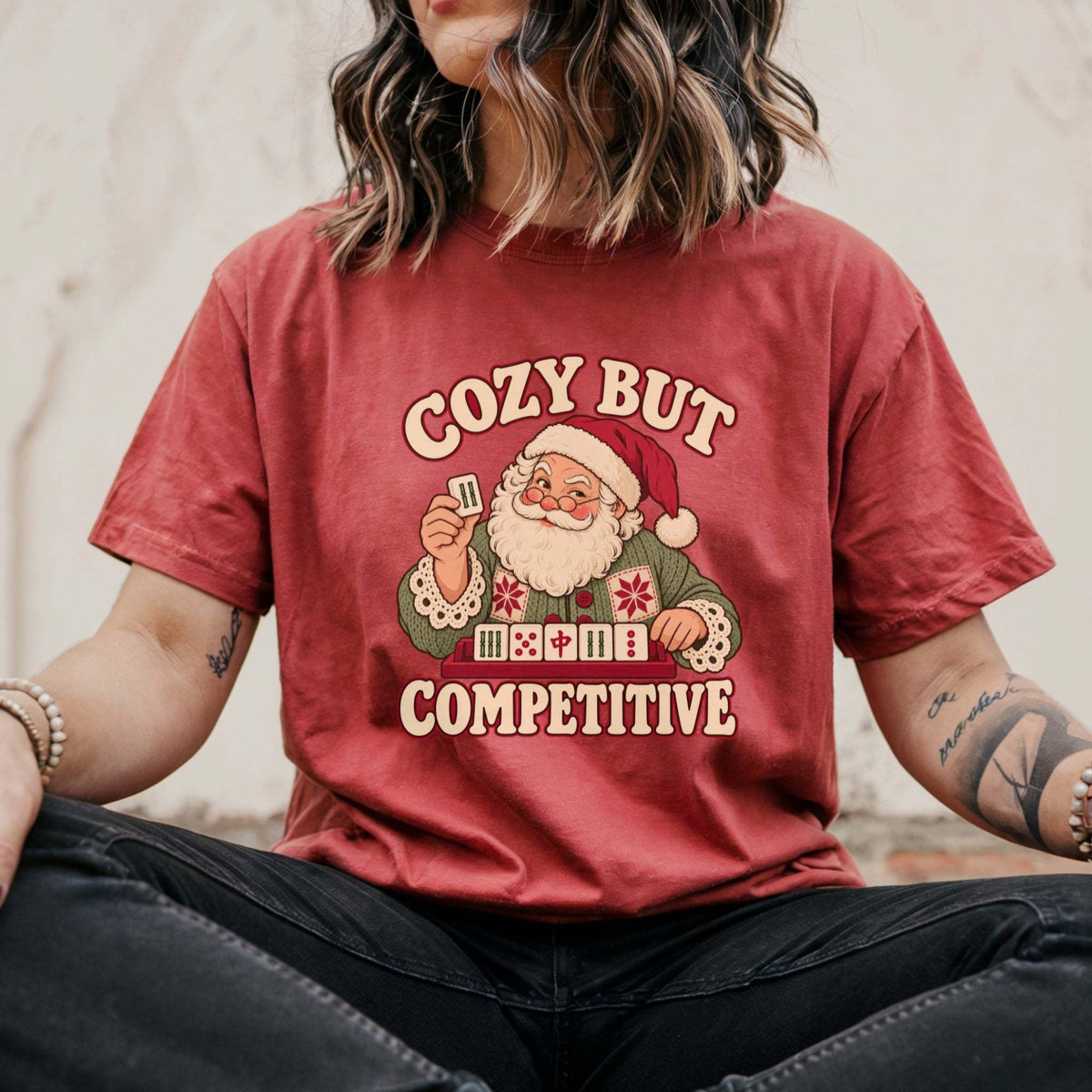 Model wearing a Crimson red Comfort Colors T-shirt with Santa holding a mahjong tile above a tile rack and cream “Cozy But Competitive” lettering.