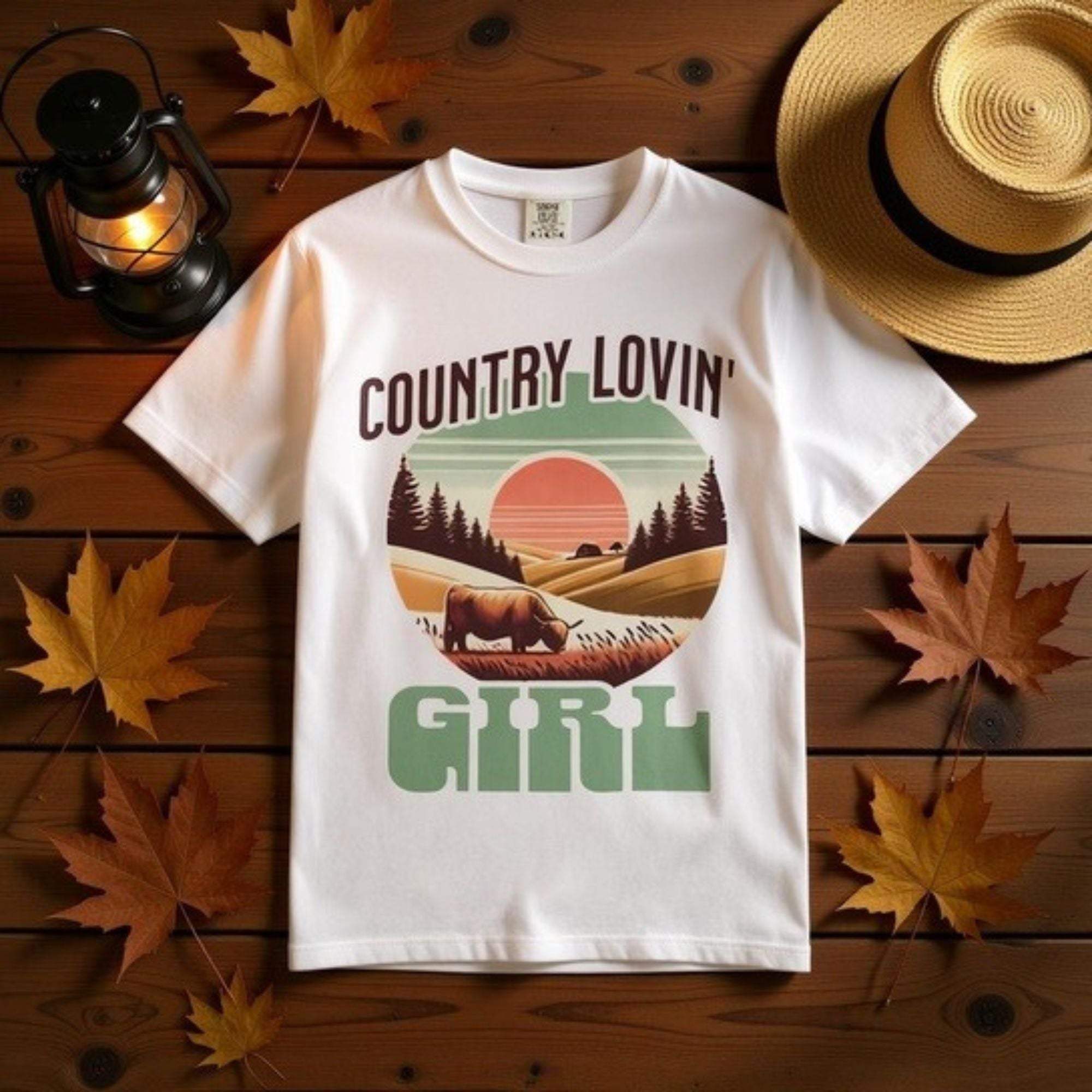 White Country Lovin Girl T-shirt with vintage rural landscape and cow design on wooden background with autumn leaves and straw hat