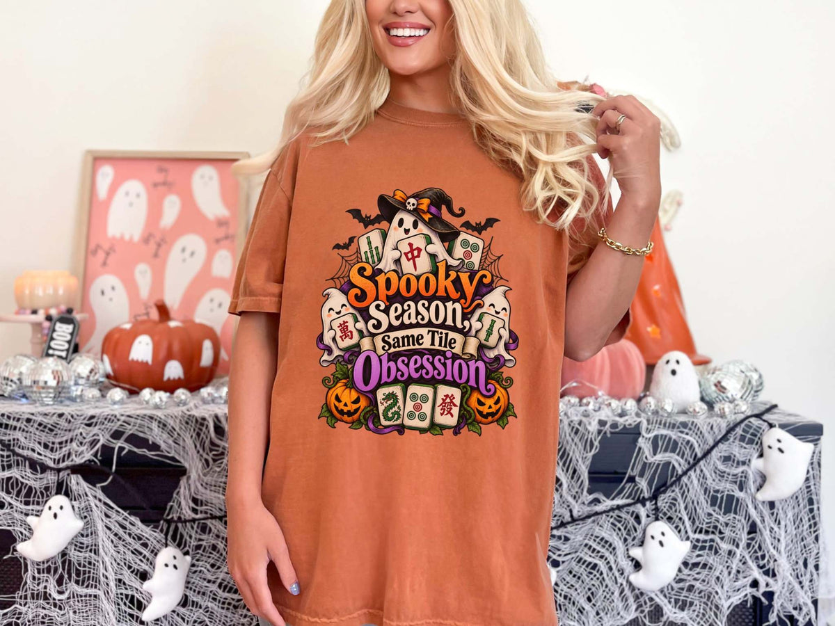 Model wearing a yam orange Halloween mahjong T-shirt with ghosts, pumpkins, mahjong tiles and “Spooky Season, Same Tile Obsession” lettering.