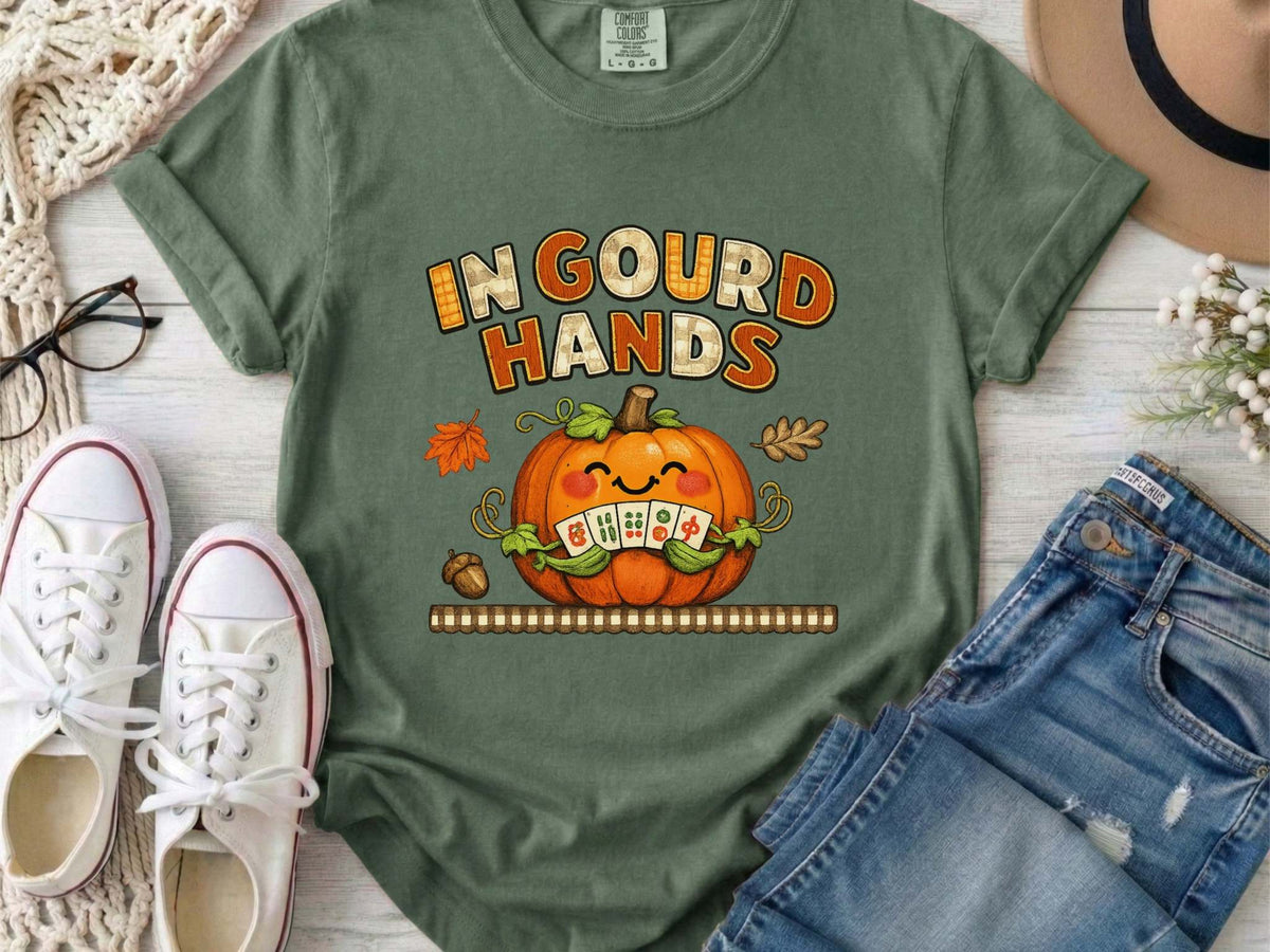 A moss green short-sleeve t-shirt with a colorful graphic on the front. The graphic features the text 'IN GOUD HANDS' in a stylized orange and brown font above a smiling orange pumpkin with a toothy grin and autumn-themed details.