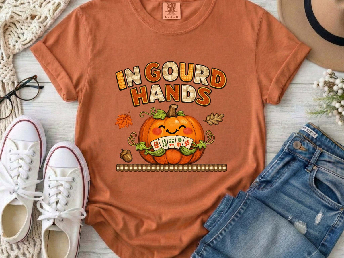 A yam orange short-sleeve t-shirt with a colorful graphic on the front. The graphic features the text 'IN GOUD HANDS' in a stylized orange and brown font above a smiling orange pumpkin with a toothy grin and autumn-themed details.