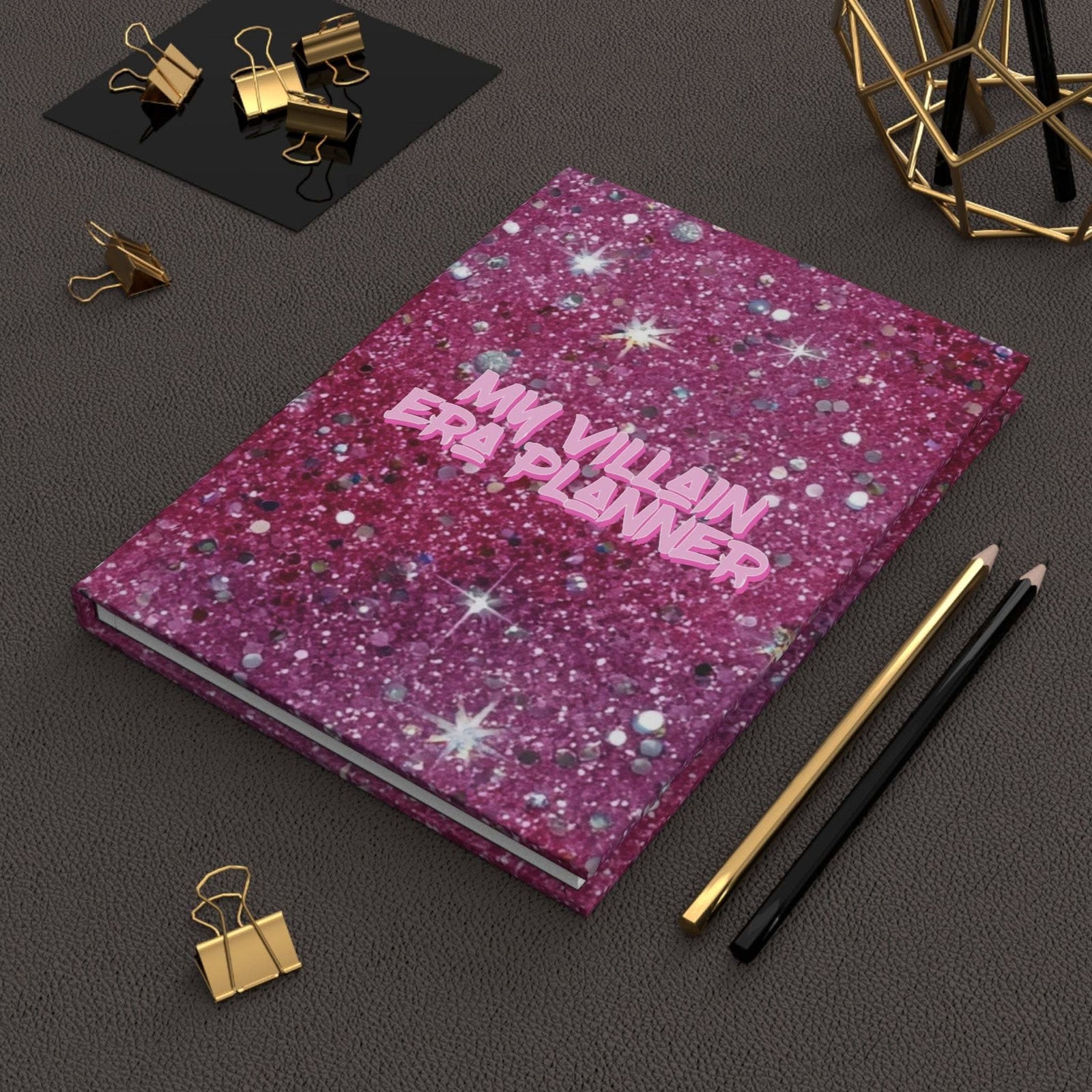 Matte hardcover journal with glittery purple cover and "My Villain Era Planner" text on dark textured surface with pencils and gold binder clips.