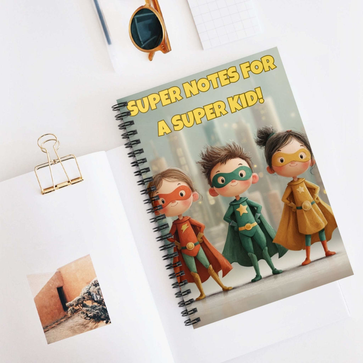 Spiral notebook with superhero theme titled Super Notes for a Super Kid on a white desk with sunglasses and paper clip