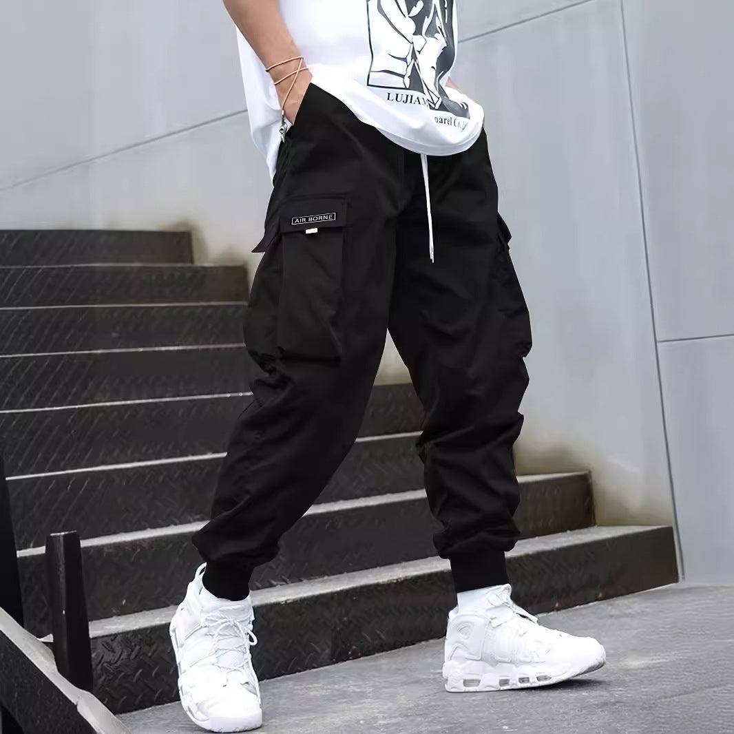 Men's black multi-pocket cargo pants with drawstring waist, casual hip-hop style, paired with white sneakers.