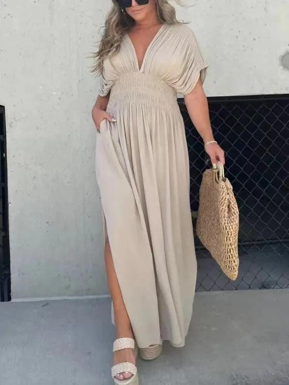 Woman wearing a beige bat-sleeved V-neck slit dress with elastic waist and short sleeves, holding a woven bag outdoors.