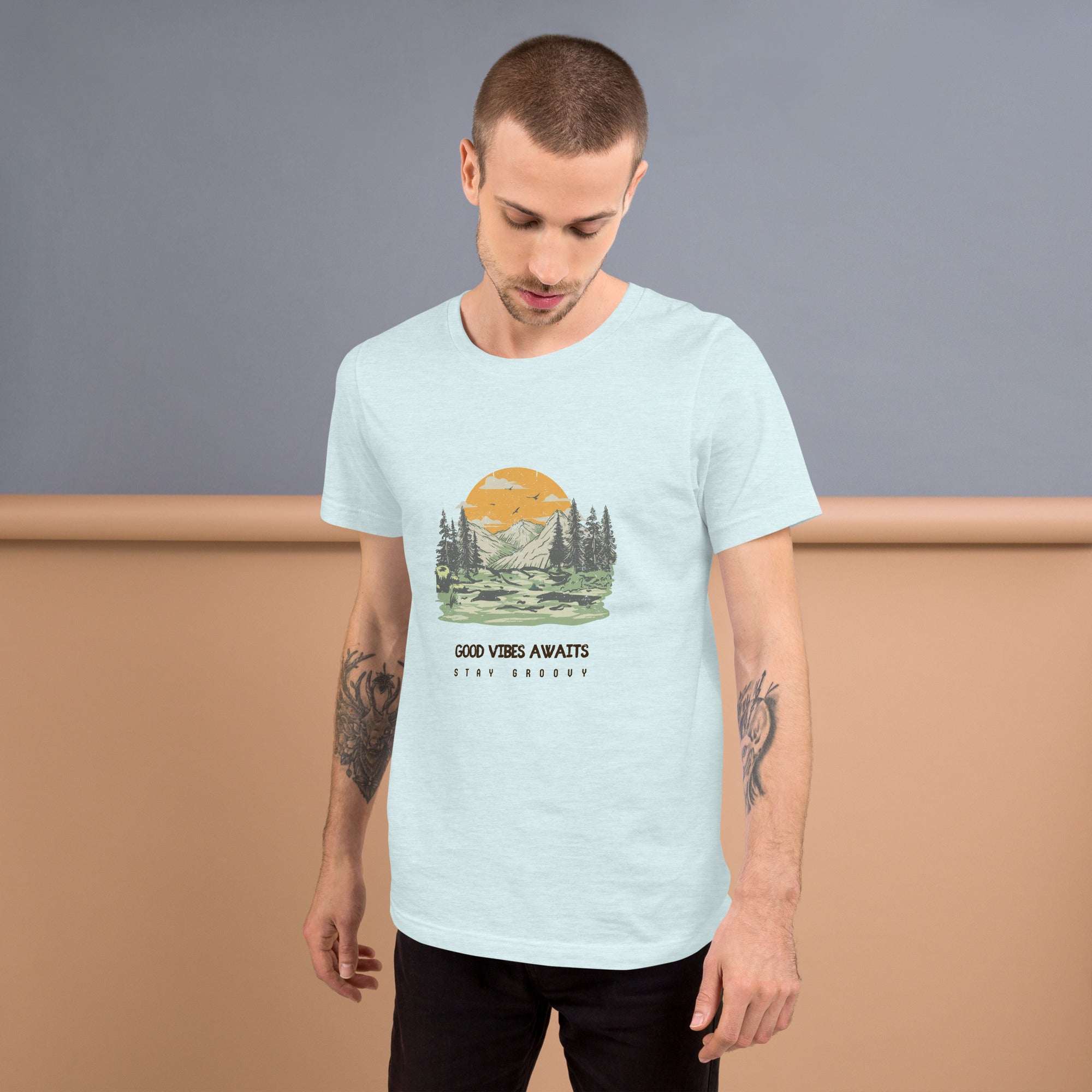 Man wearing light blue Good Vibes Awaits T-Shirt with nature graphic and text, standing against gray and beige background