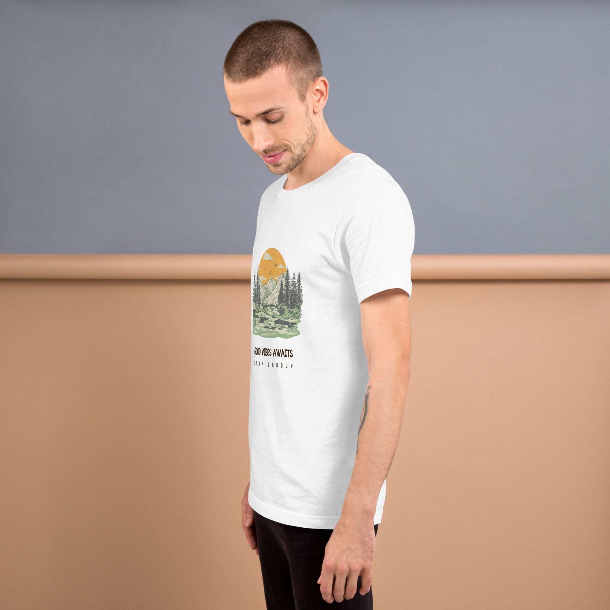 Man wearing white Good Vibes Awaits T-Shirt with nature graphic and text on front, posing against gray and beige background