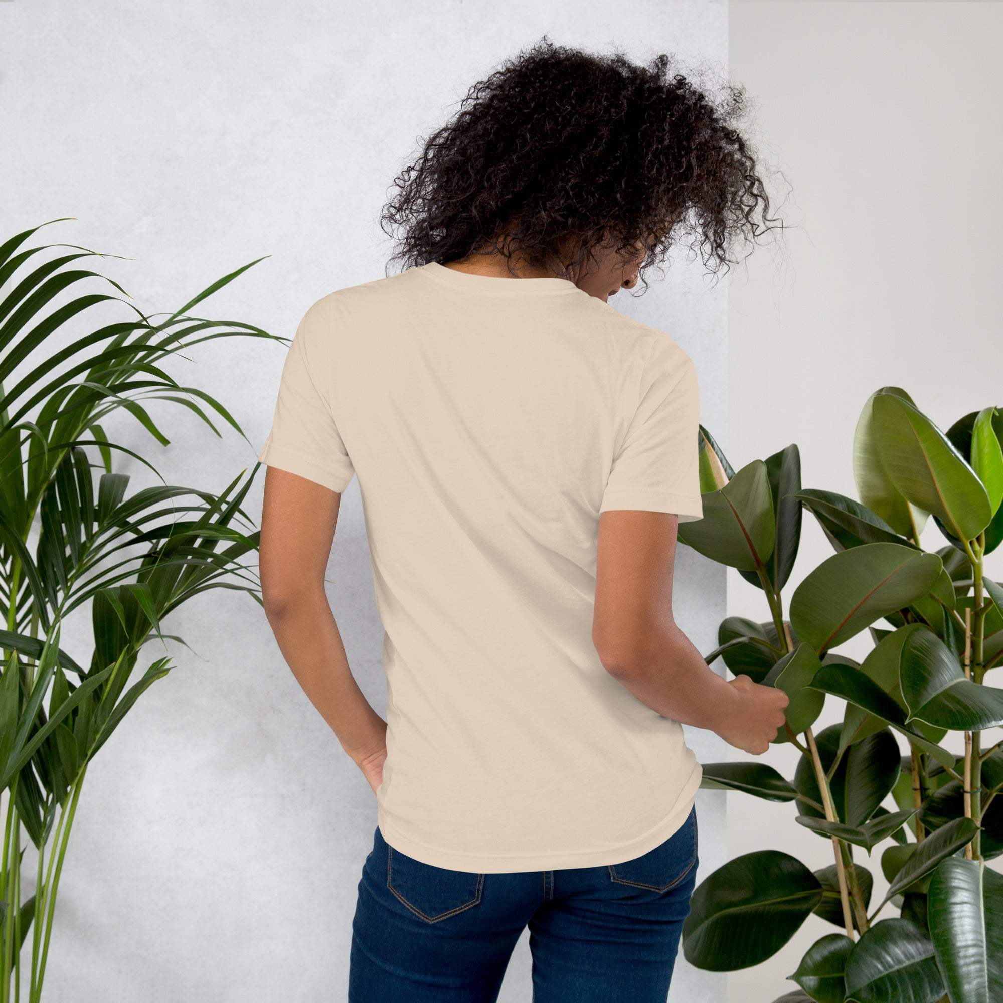 Woman wearing beige coffee t-shirt standing among green plants showing back design