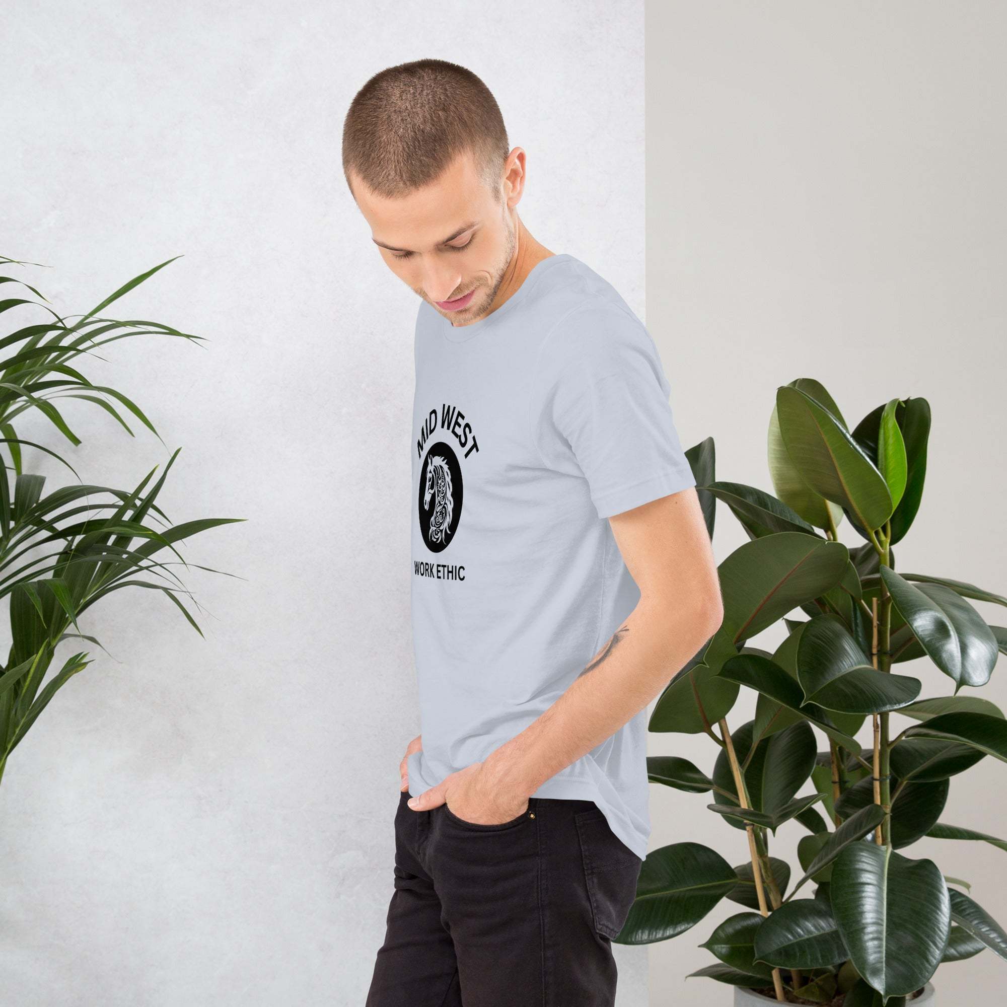Man wearing light gray Midwest T-Shirt with horse graphic and work ethic text standing indoors near plants