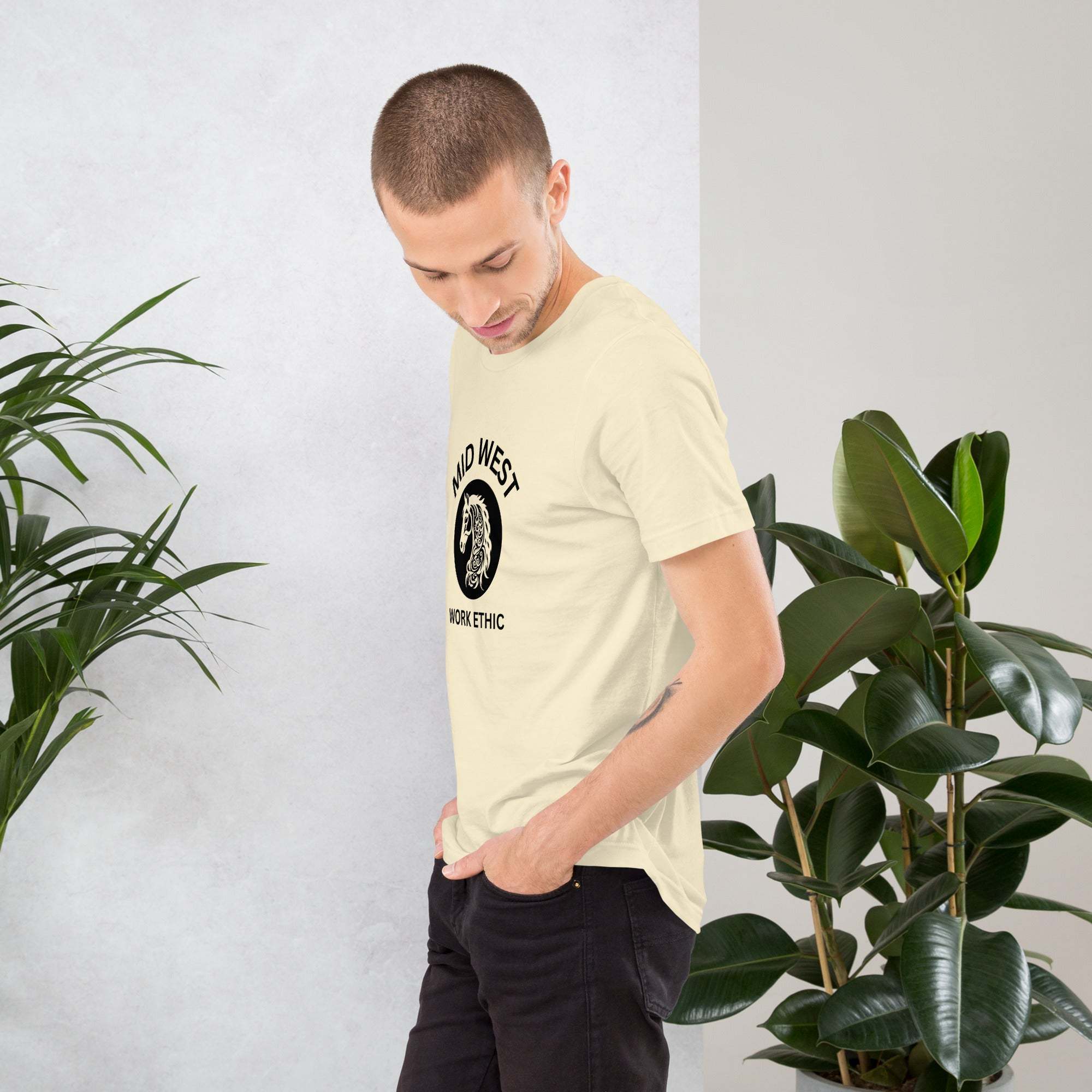 Man wearing a beige Midwest T-Shirt with horse graphic and work ethic text standing near plants
