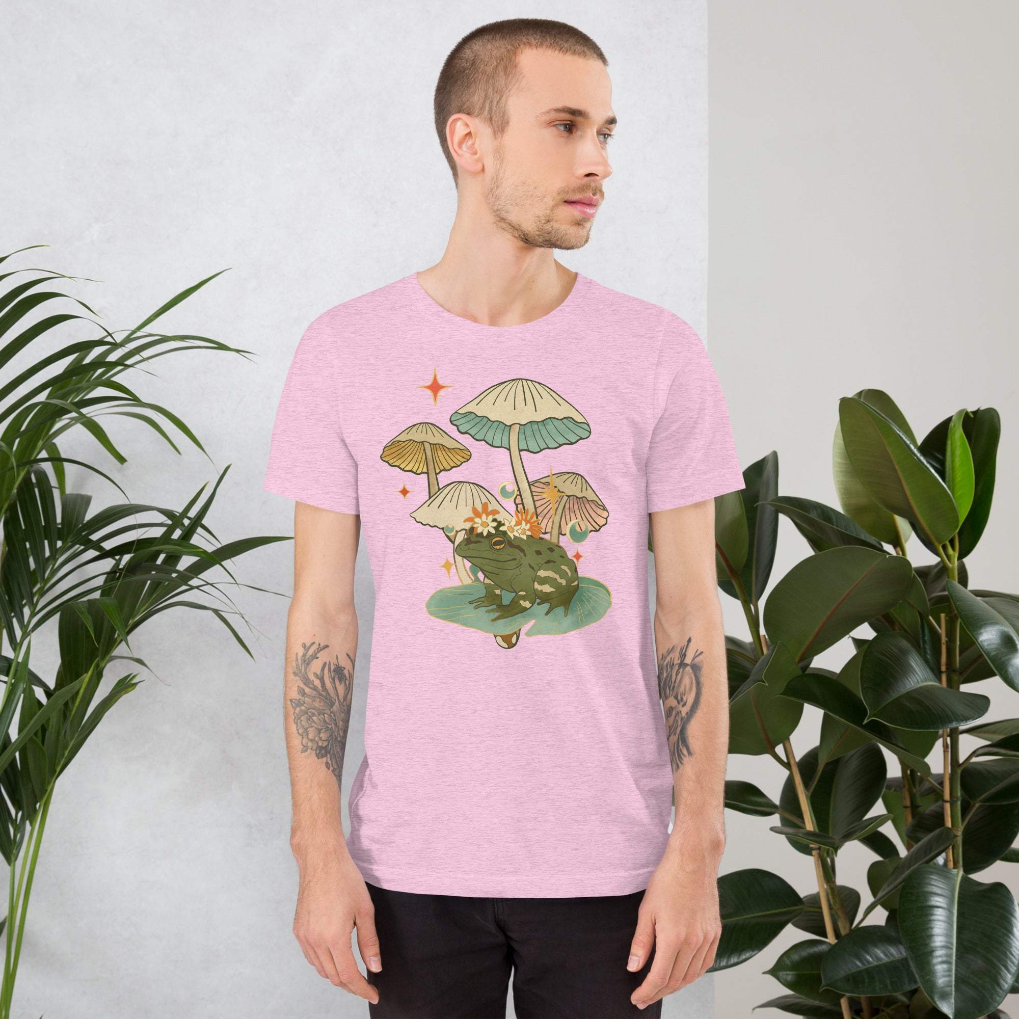 Man wearing a pink vintage aesthetic frog and mushroom t-shirt with cottagecore toadstool graphic design surrounded by green plants