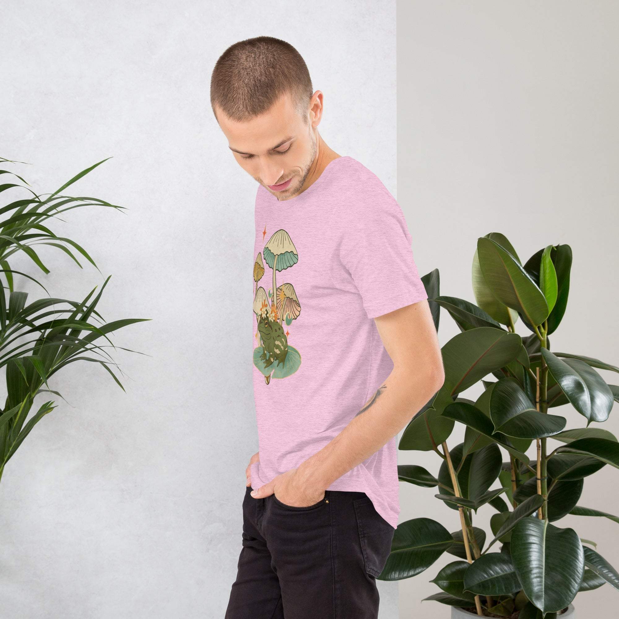 Man wearing pink vintage aesthetic frog and mushroom T-shirt standing beside green plants