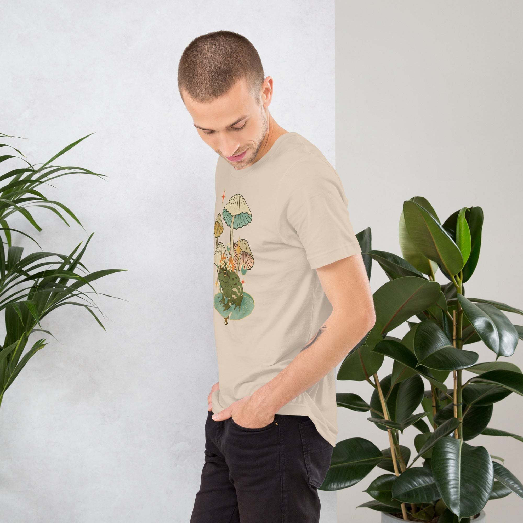 Man wearing vintage aesthetic frog and mushroom graphic t-shirt standing near green plants indoors