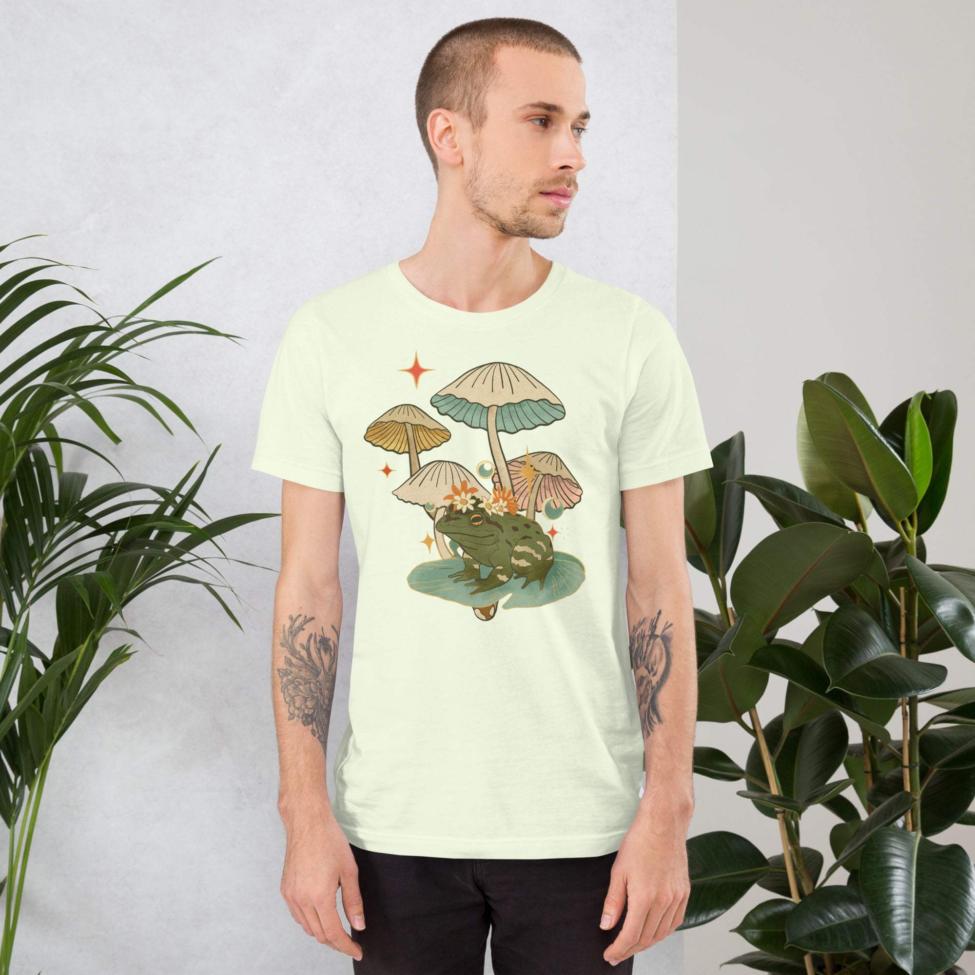 Man wearing vintage aesthetic frog and mushroom t-shirt with cottagecore graphic design standing near plants
