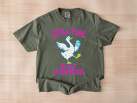 A moss green Comfort Colors crewneck t-shirt featuring a graphic of a white goose holding a blue water bottle, with the pink text 'STILL FUN. JUST HYDRATED.