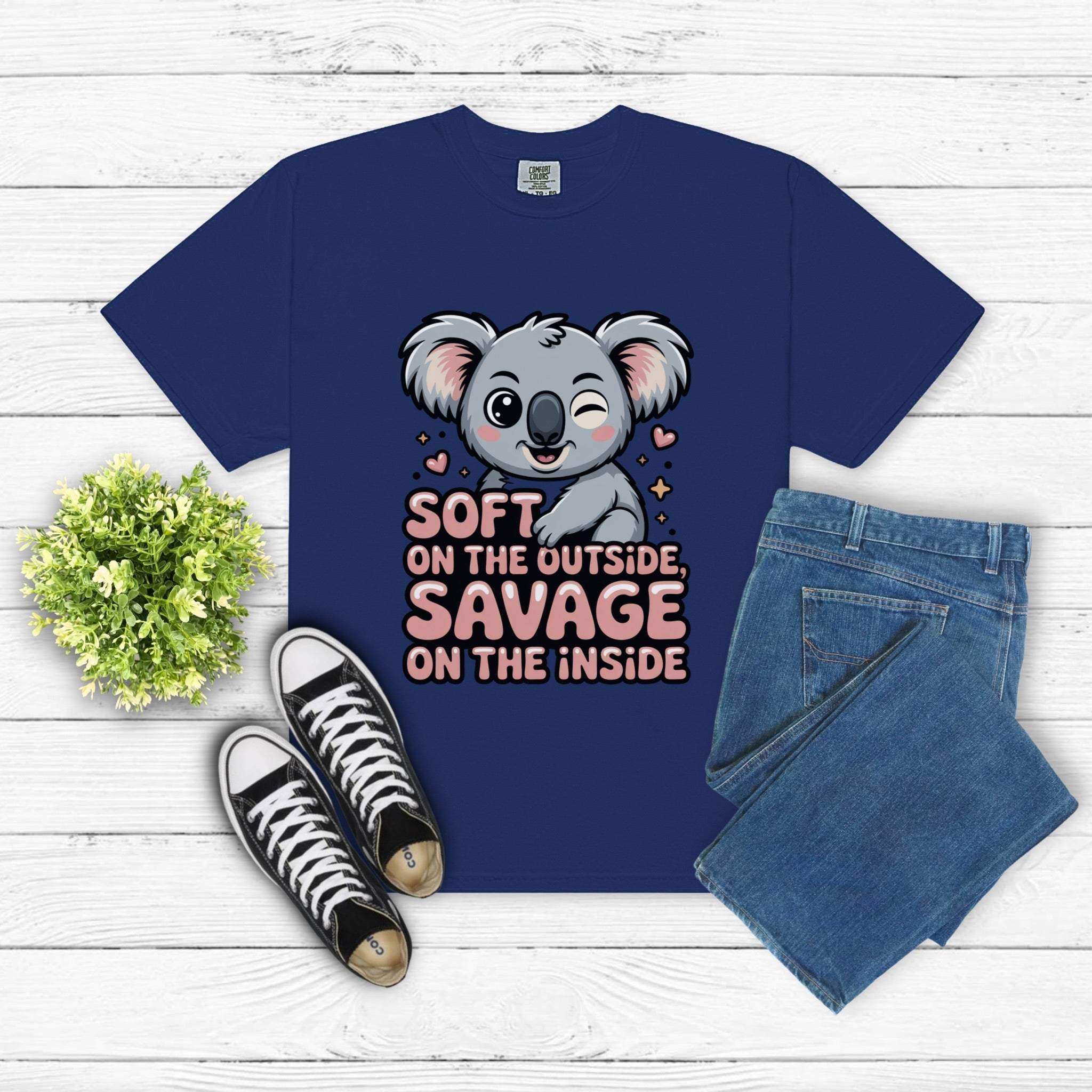 Blue t-shirt with cute koala and "Soft on the Outside Savage on the Inside" text design laid out with jeans and sneakers on white wooden background