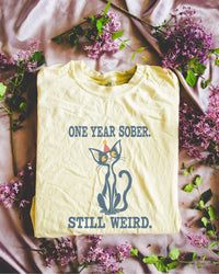 Folded flat lay of a butter Comfort Colors 1717 t-shirt featuring a navy cat in a red party hat and “One Year Sober. Still Weird.” lettering.