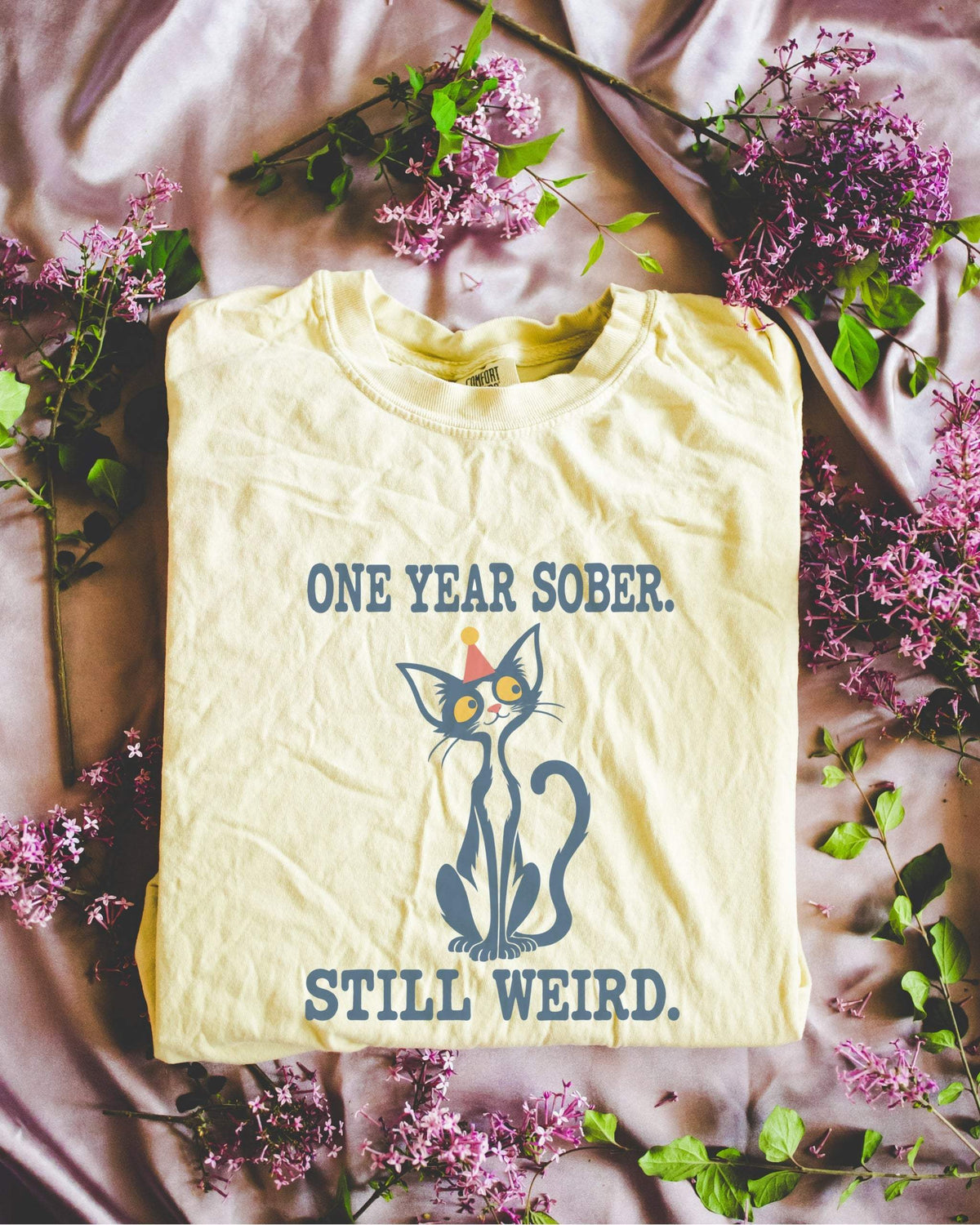 Folded flat lay of a butter Comfort Colors 1717 t-shirt featuring a navy cat in a red party hat and “One Year Sober. Still Weird.” lettering.