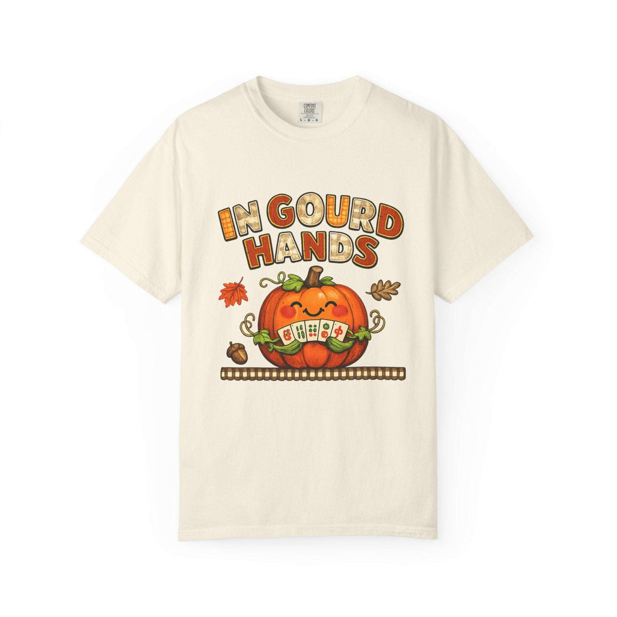 An off-white short-sleeve t-shirt with a colorful graphic on the front. The graphic features the text 'IN GOUD HANDS' in a stylized orange and brown font above a smiling orange pumpkin with a toothy grin and autumn-themed details.