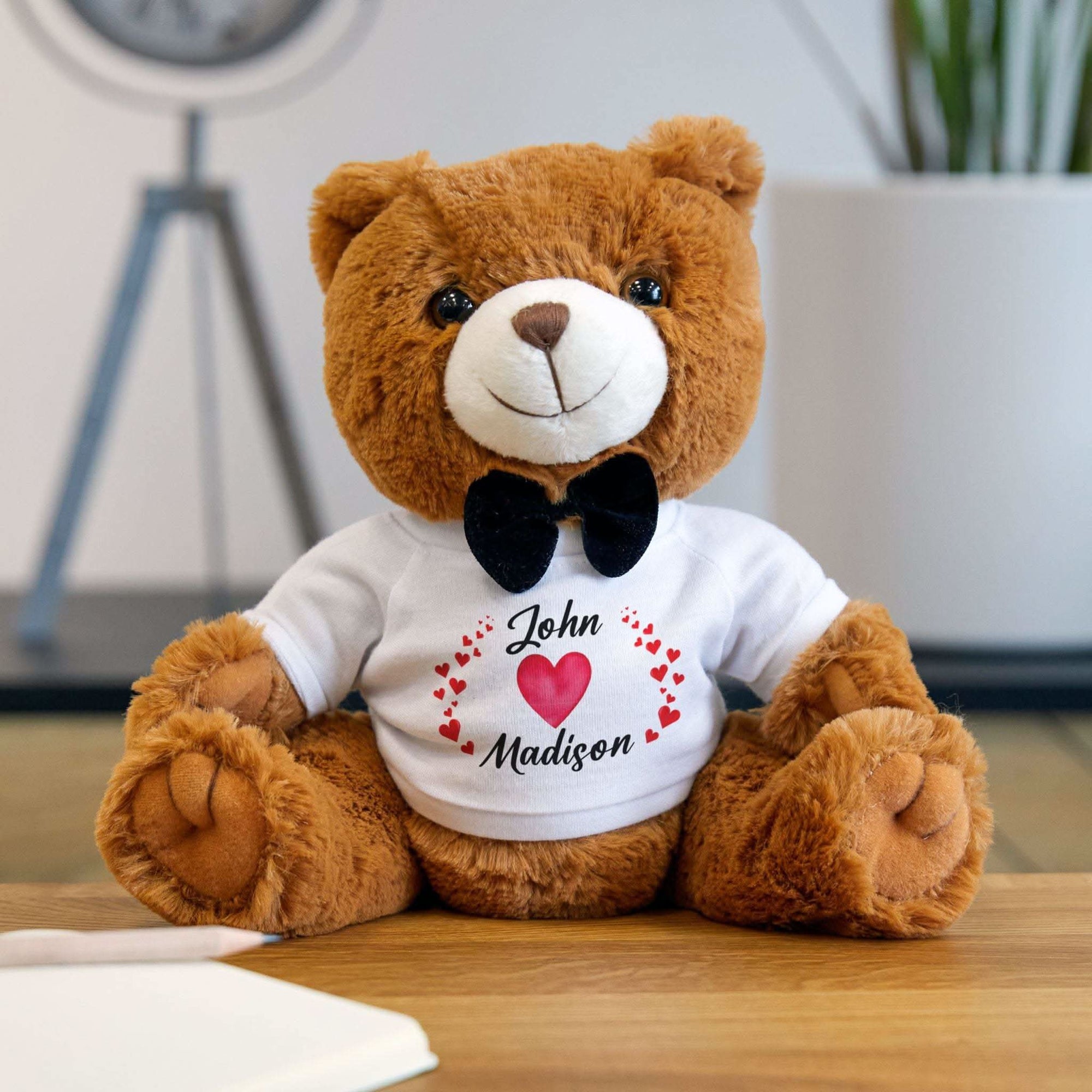 Seated light-brown teddy bear wearing a white T-shirt with personalized heart names and black bow tie, perfect Valentine's gift