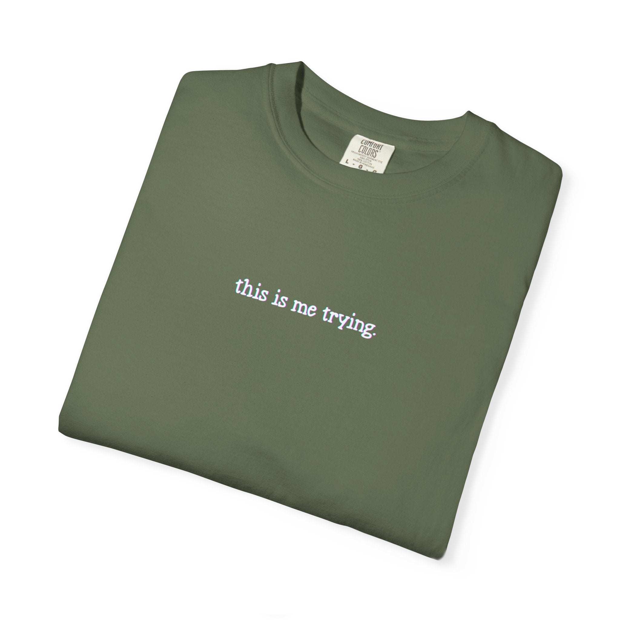 Folded green t-shirt with 'this is me trying' text on a white background