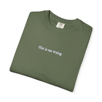 Folded green t-shirt with 'this is me trying' text on a white background