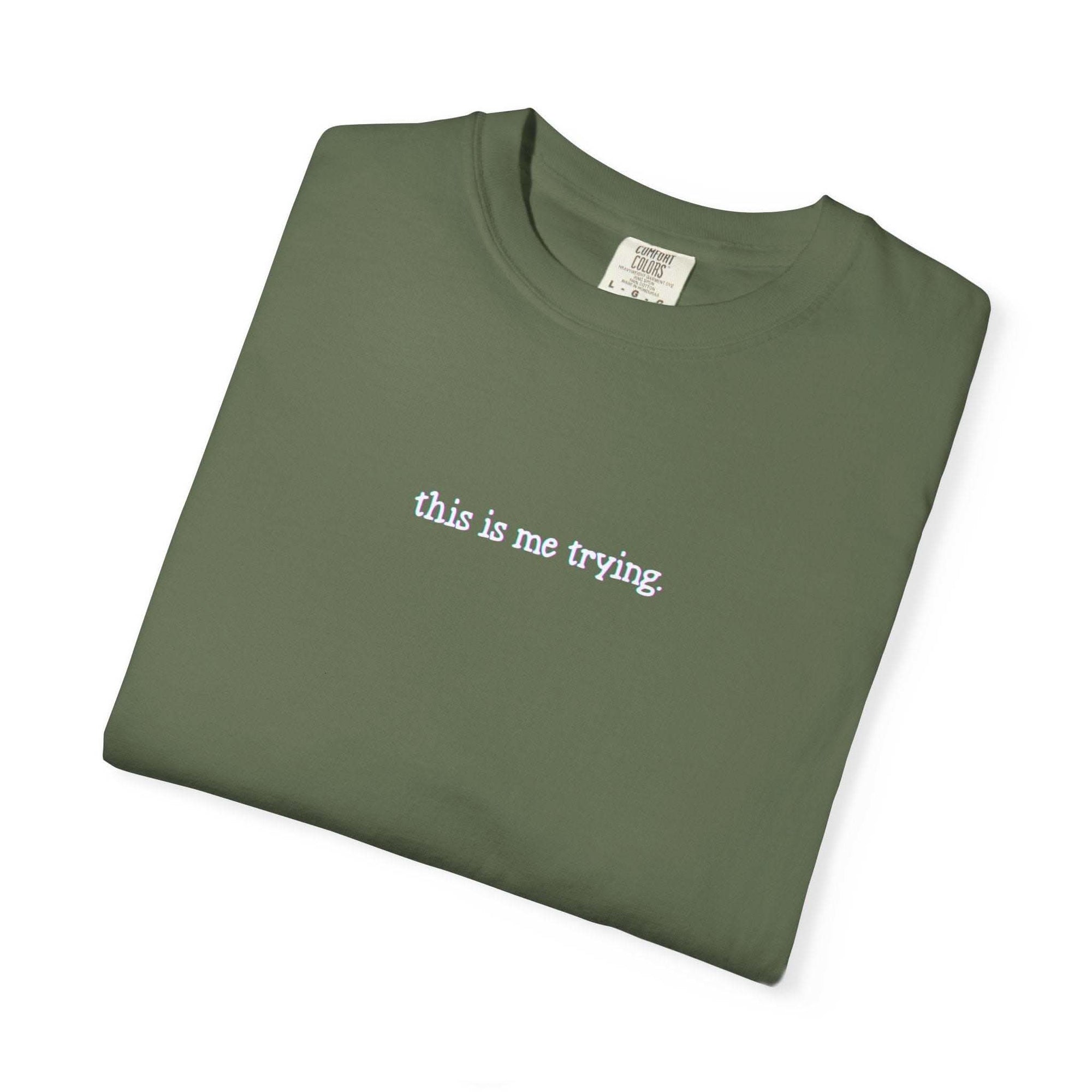 Folded green t-shirt with 'this is me trying' text on a white background