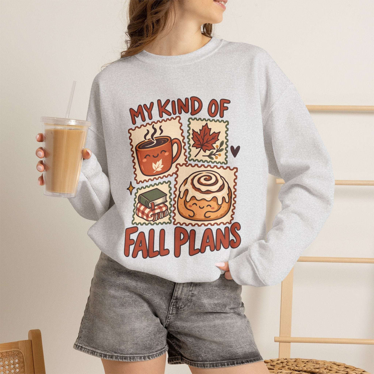 A woman wearing a light gray crewneck sweatshirt with a colorful graphic that reads 'MY KIND OF FALL PLANS' and showing illustrations of a coffee mug, autumn leaf, candy corn, and cinnamon roll. She is holding an iced coffee and wearing denim shorts in a warm indoor setting.